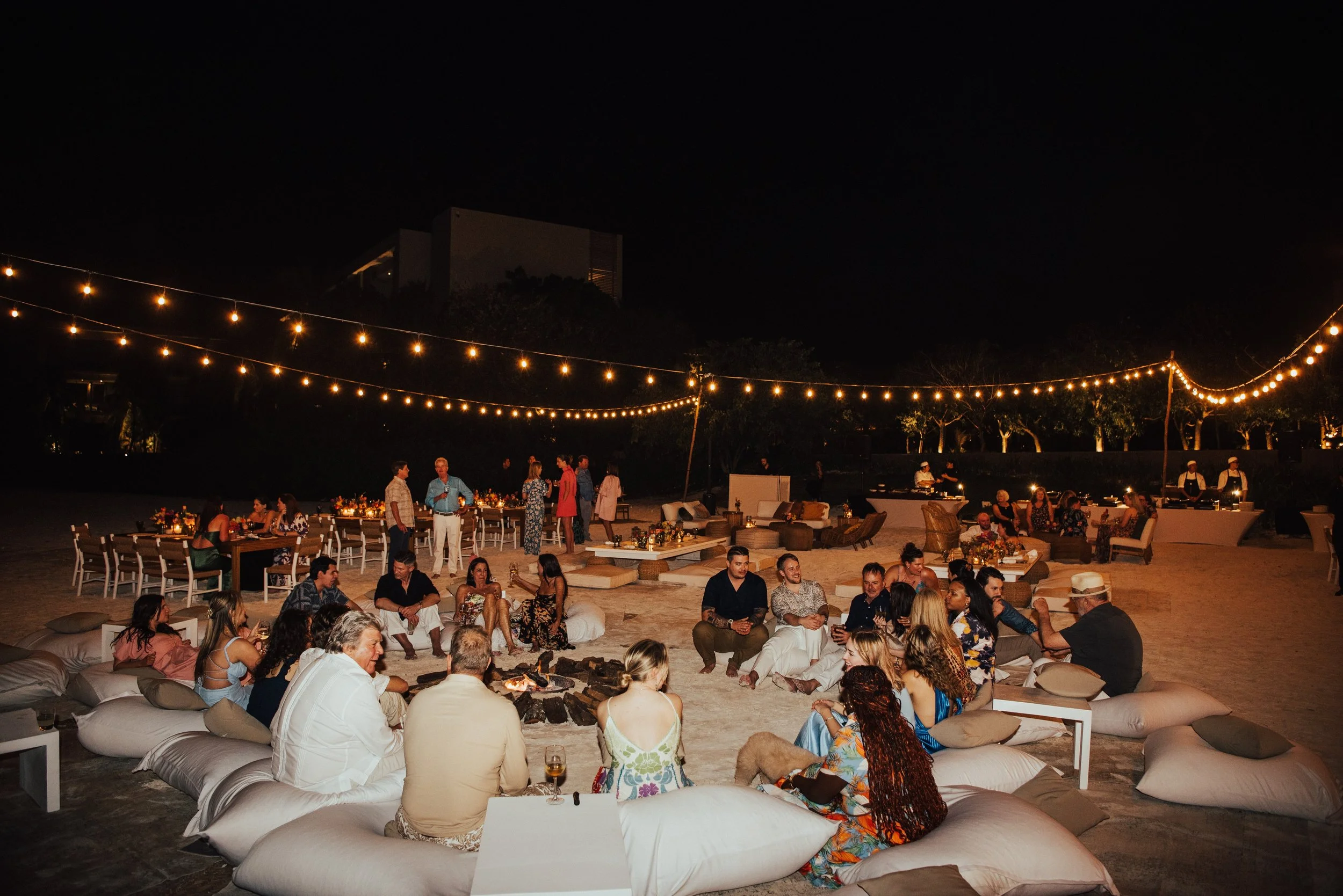 wide shot of wedding welcome party on the beach at night. String lights over the guest as they sit around a fire on cushions.