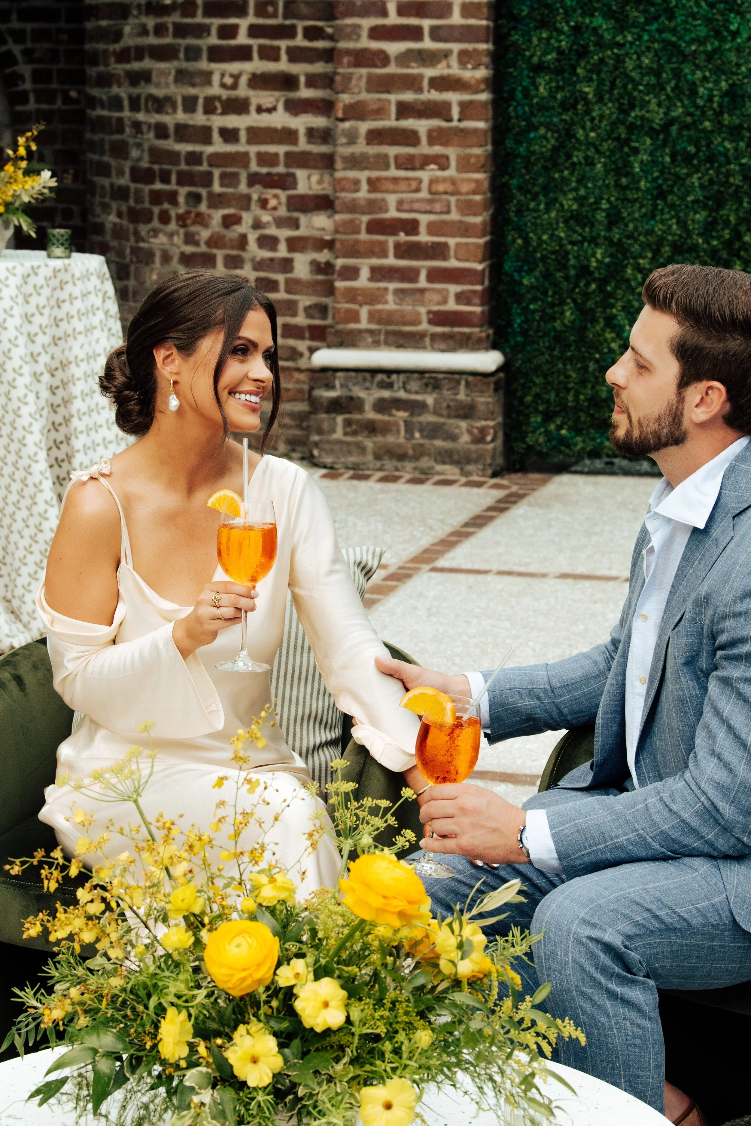 woman in white dress smiling at man in blue suit as they sit with orange drinks in their hands
