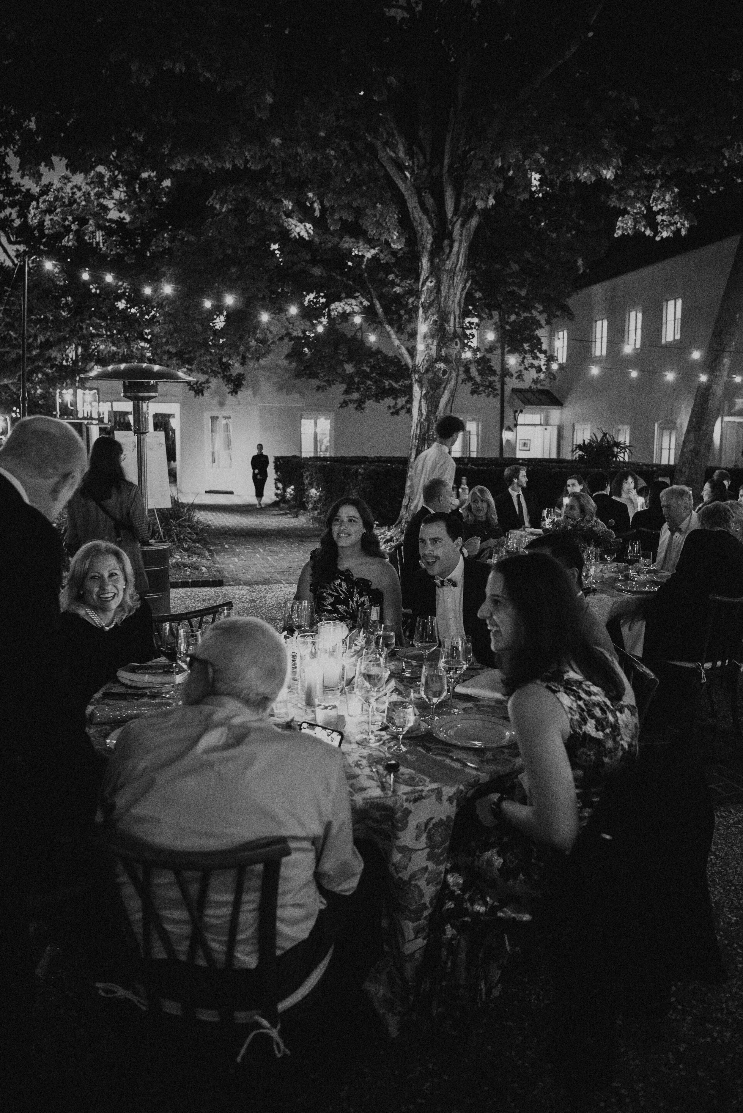 Black and white photo of guest laughing around a round table at night under bistro lights