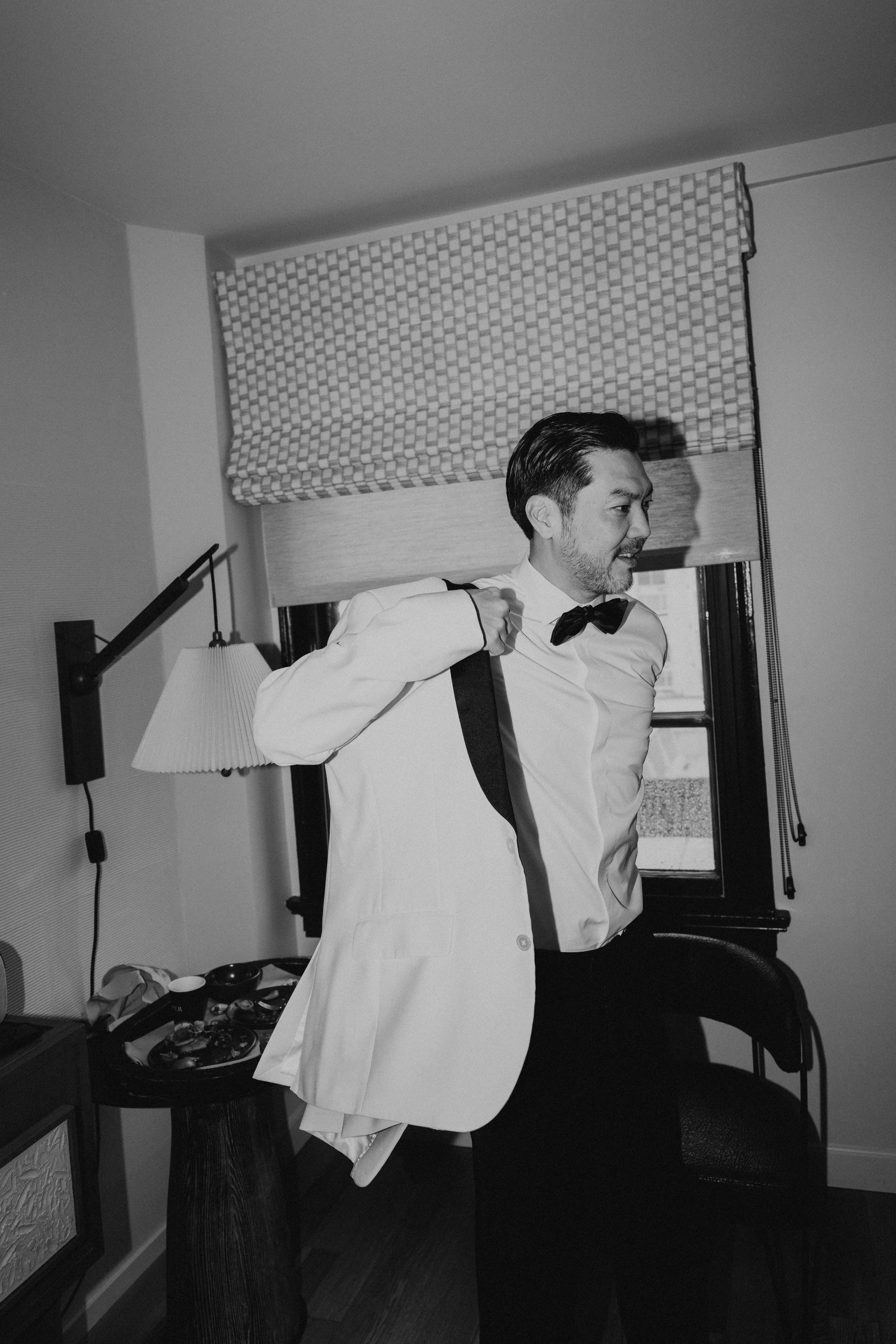 Black and white photo of groom putting on his white tuxedo jacket