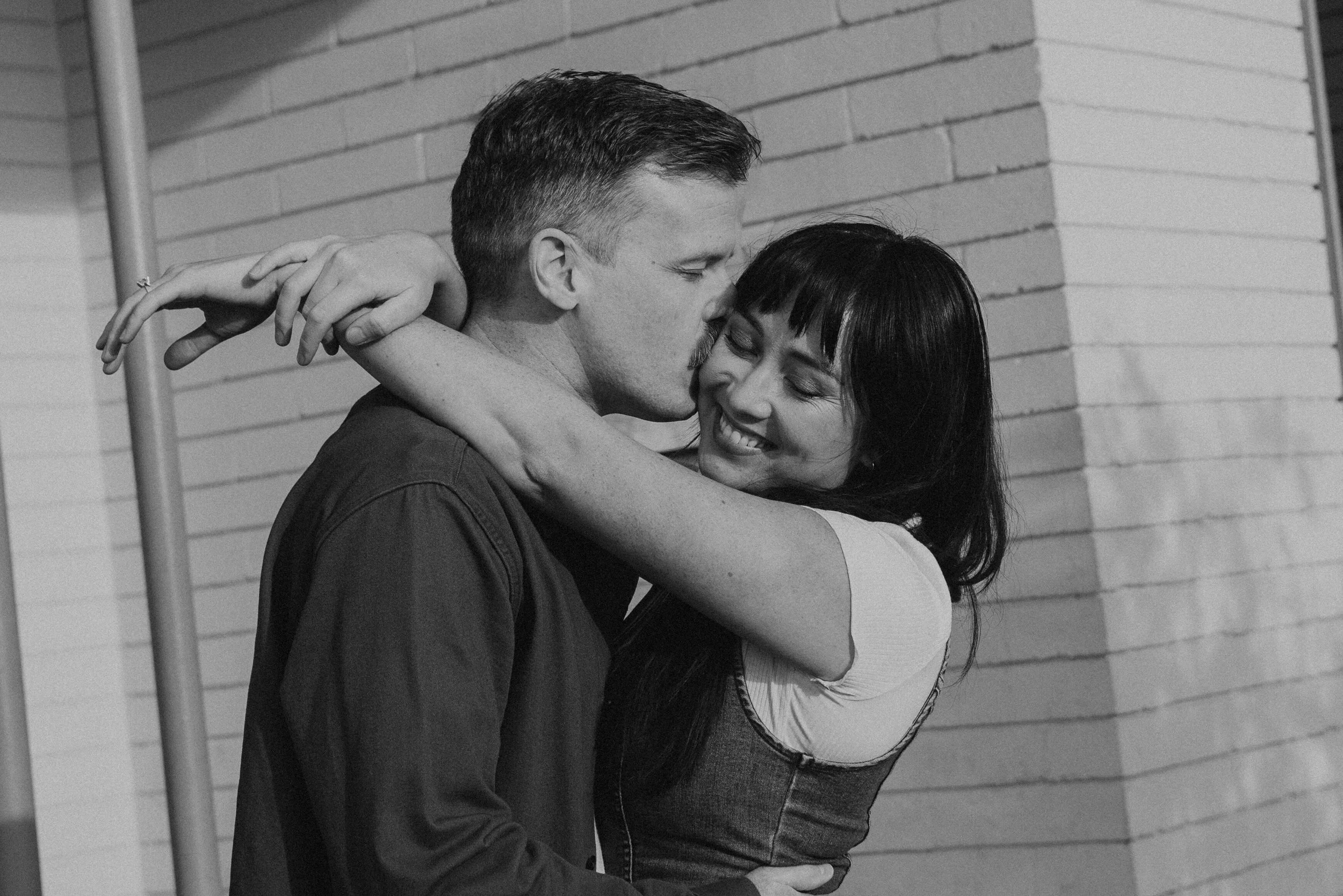 Black and white photo of guy kissing girls cheek while she smiles