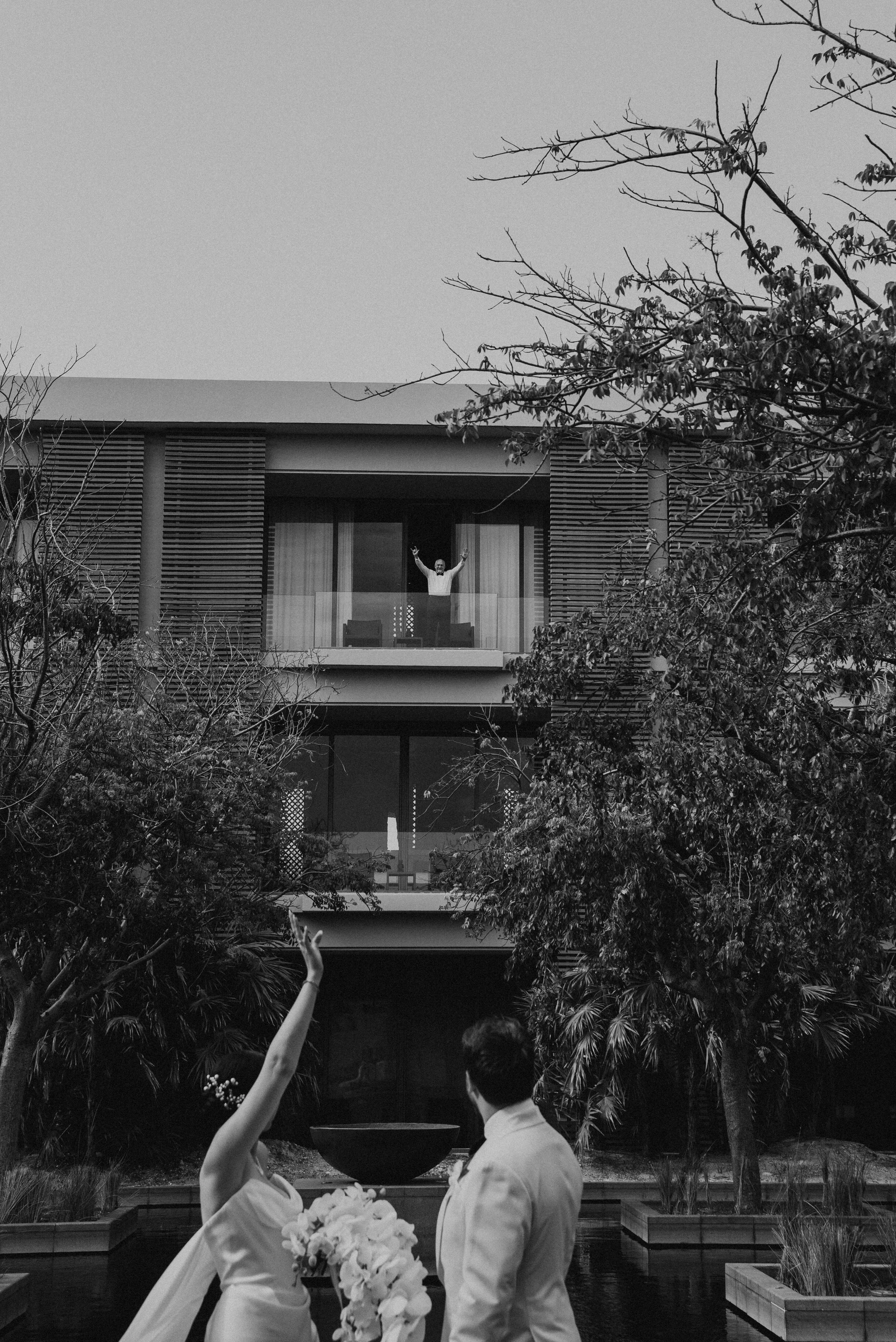 Black and white photo of bride and groom waving at man on a balcony a few stories up behind them