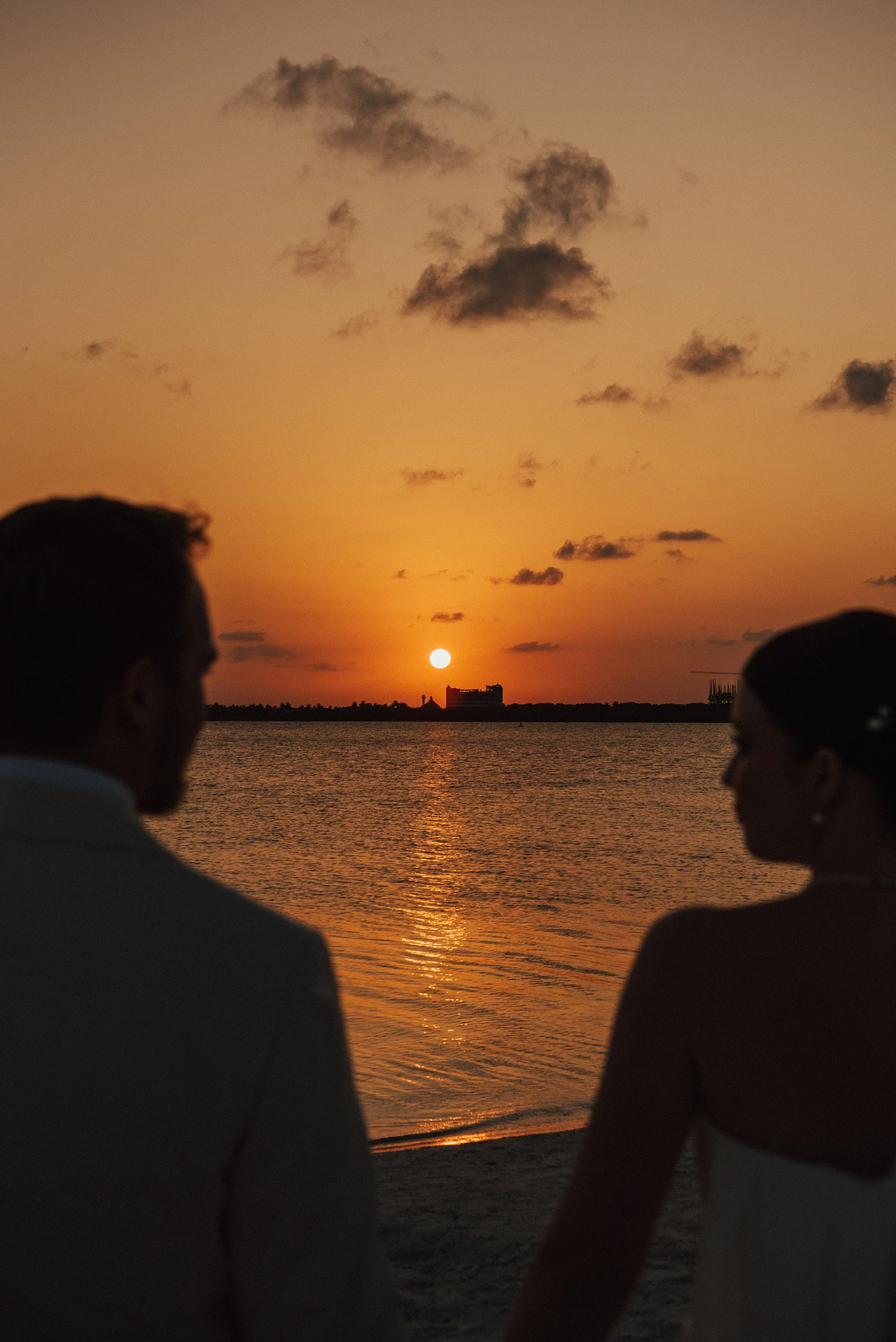 bride and groom silhouettes with heads turn towards each other. Sun can be seen setting between them
