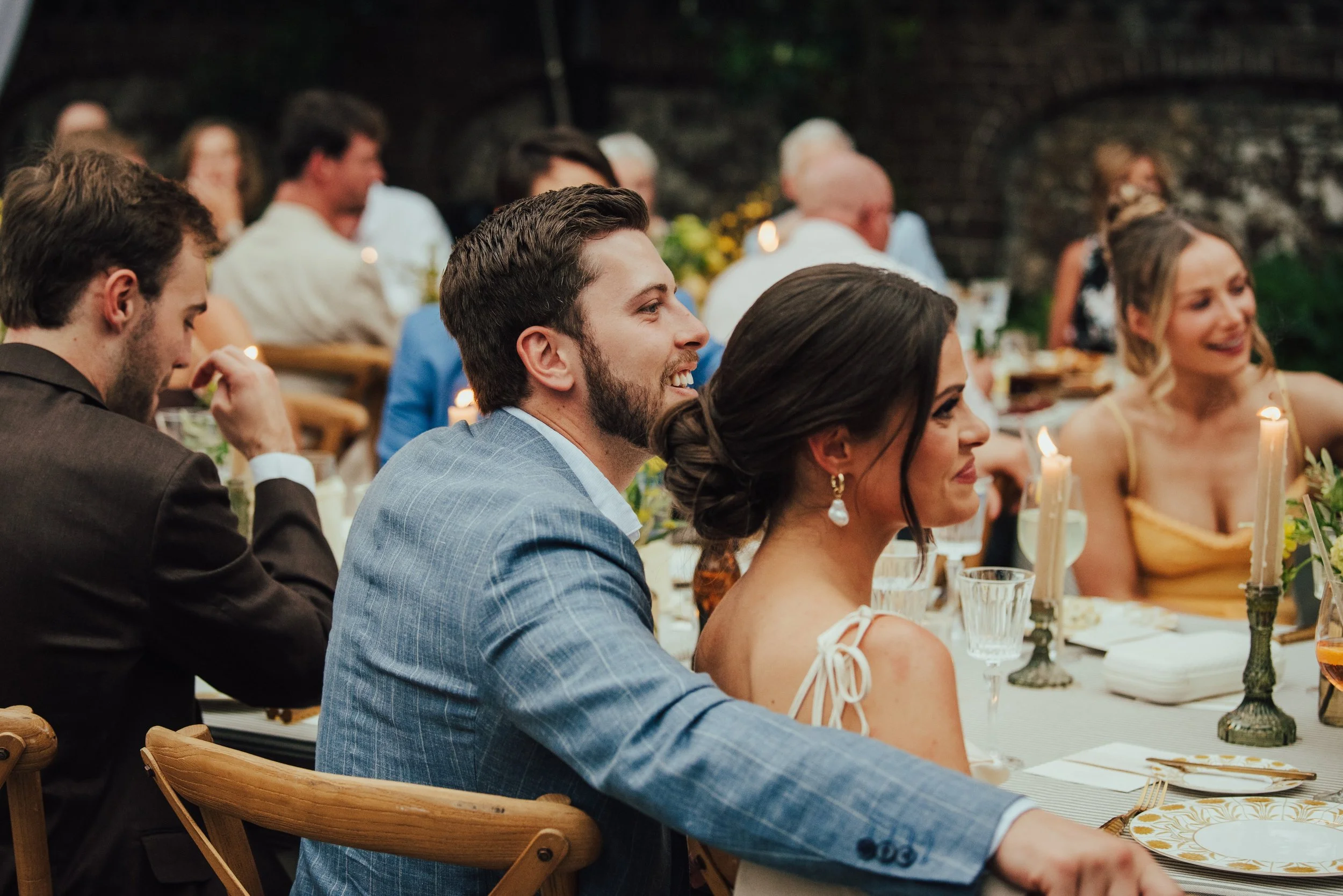 Man in blue suit with his arm on the back of a woman in white dress chair. They are listening to someone speak