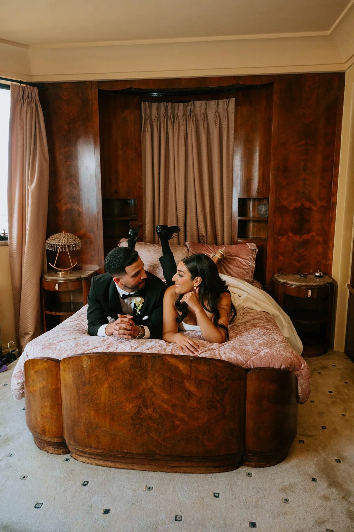 bride and Groom laying on their stomach on a pink bed pushed up on their elbow looking at each other