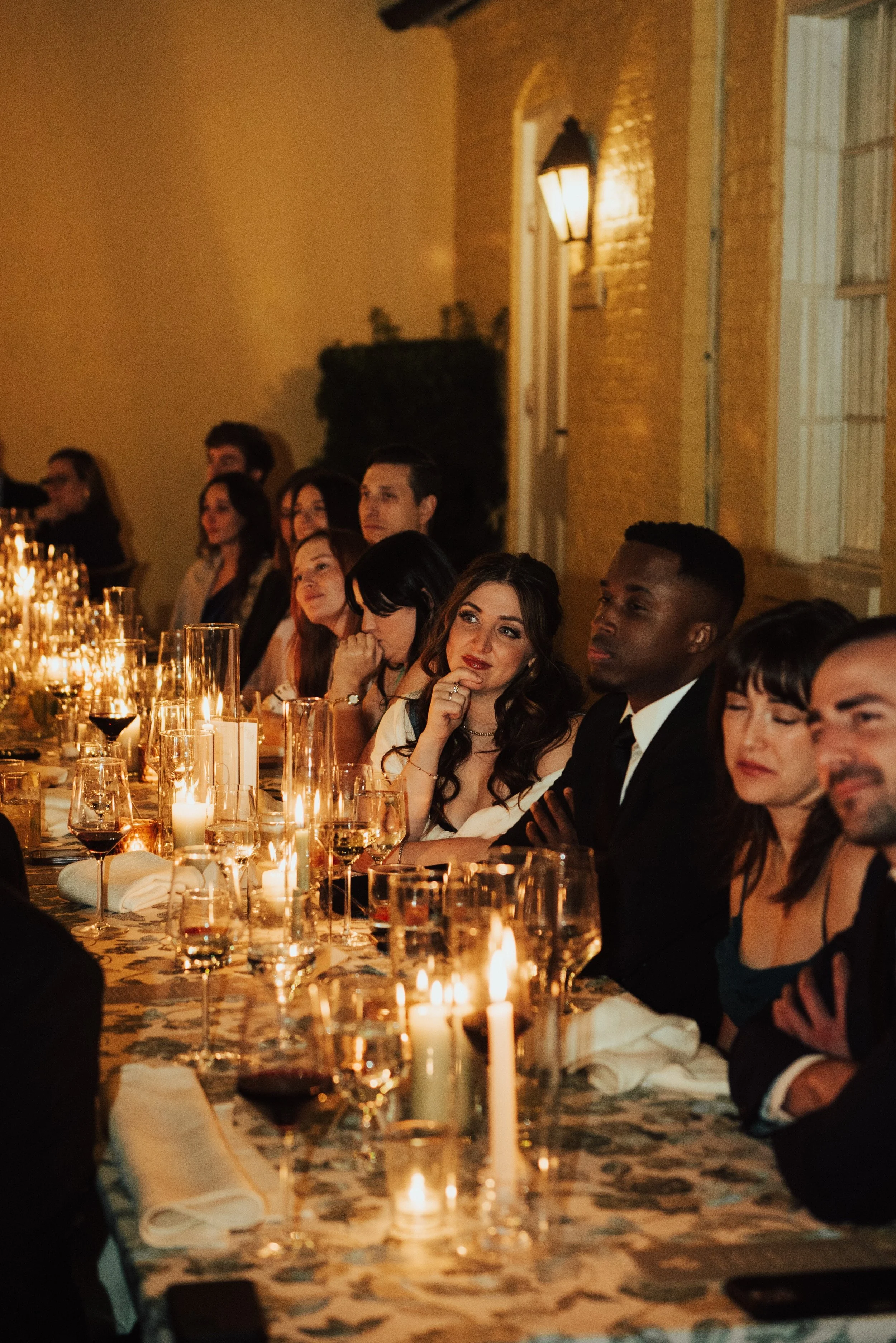 Guests sitting at a long table watching as wedding speeches take place. The table is lined with candles and wine glasses.