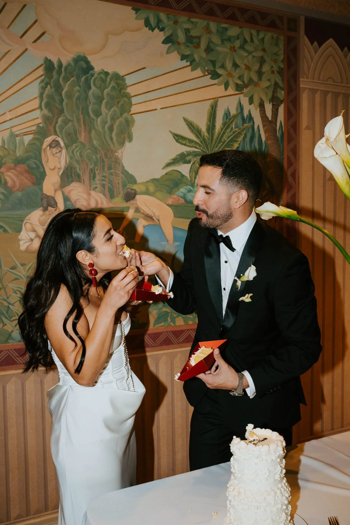 bride and groom standing in front of a mural. Groom is feeding bride cake.