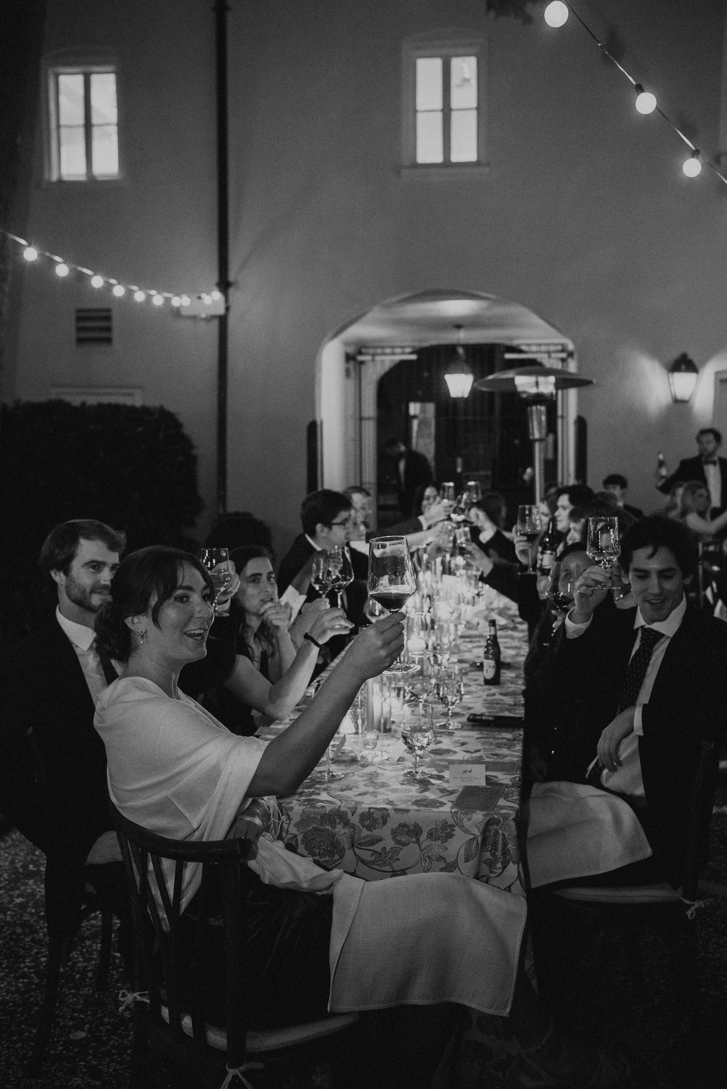 Black and white photo of guest at a long table raising glasses to toast at a wedding reception