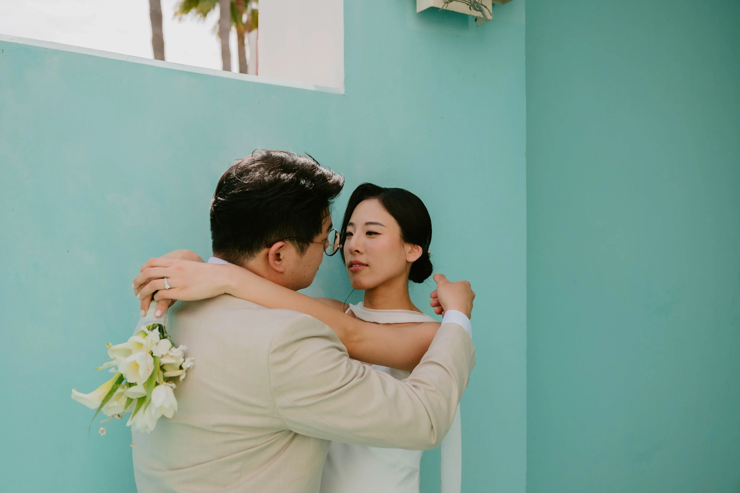 bride and Groom standing in front of a turquoise wall. Groom is fixing brides hair as she stares into eyes