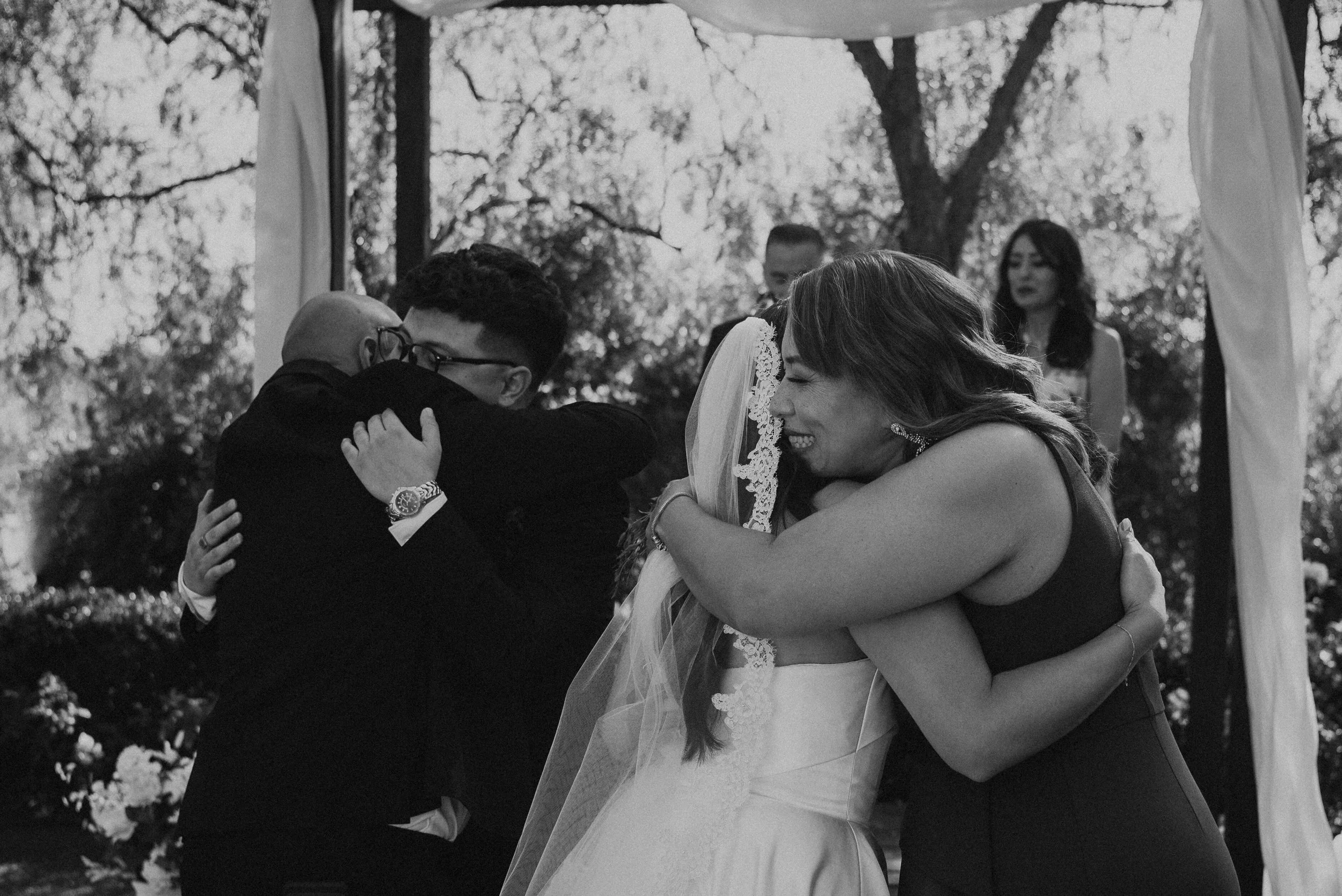 Black and white photo of bride and groom hugging her parents at the start of the ceremony
