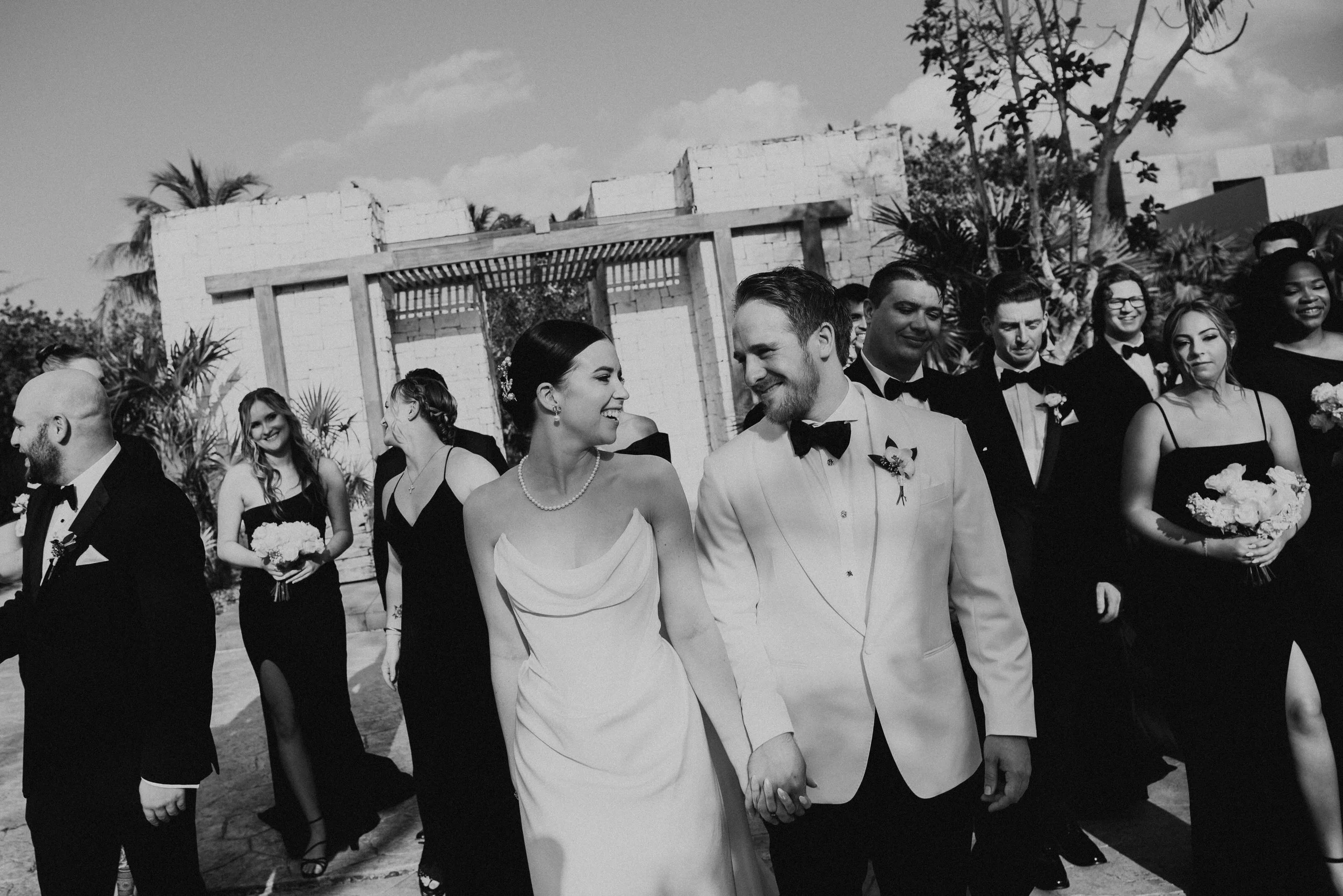 bride and groom walking towards the camera. They are holding hands smiling at each other with their wedding party behind them