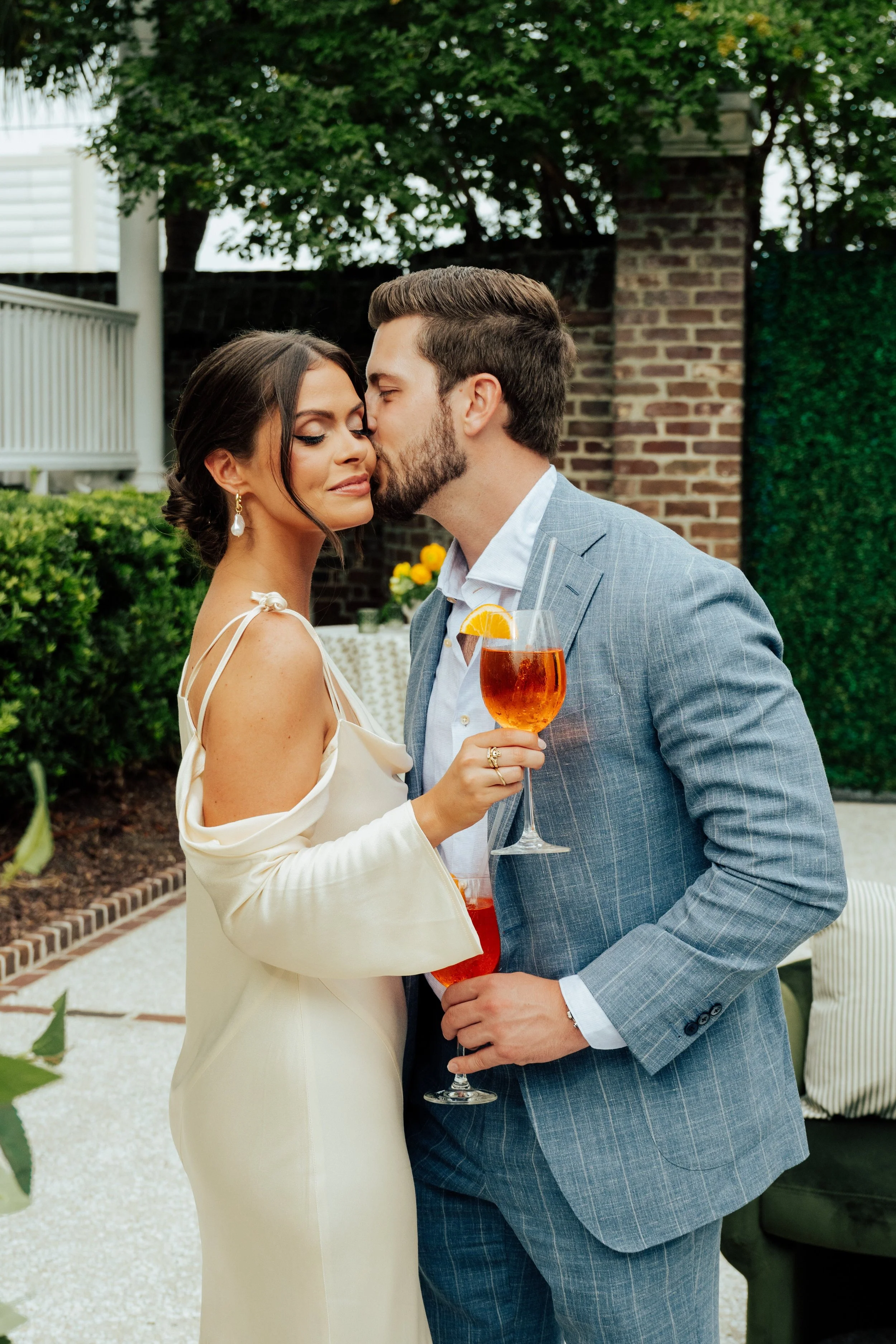 Man in blue suit kissing woman in white dress on the cheek as they hold orange drinks