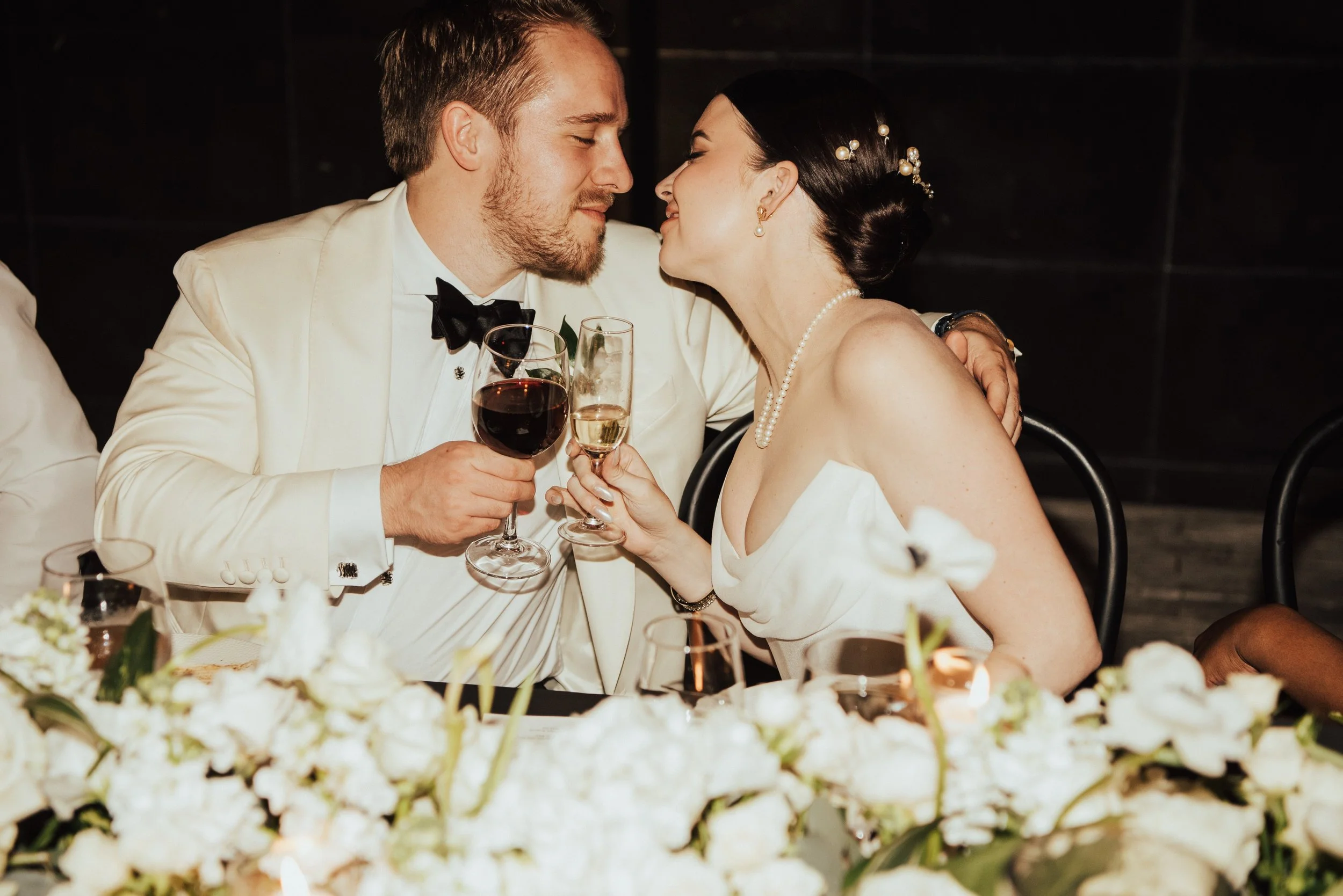 bride and groom leaning towards each other at their table clinking their glasses as they smile at each other