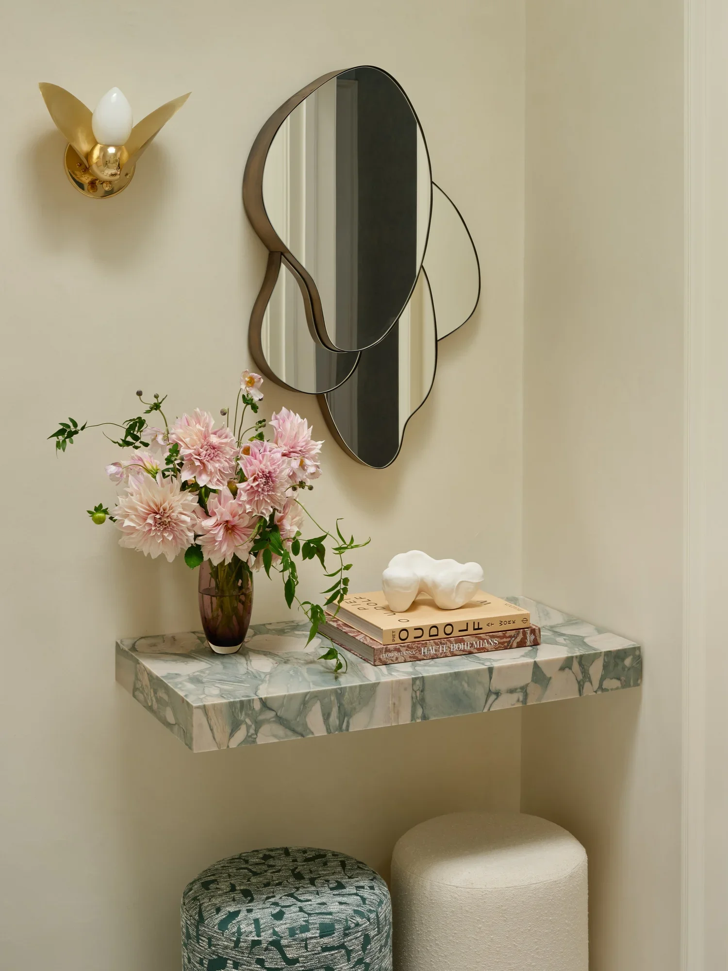 A decorative wall nook with a marble shelf, a pink flower bouquet, a stack of books, a white artistic sculpture, a small round ottoman, and a patterned ottoman. A mirror and a wall sconce are on the wall above.