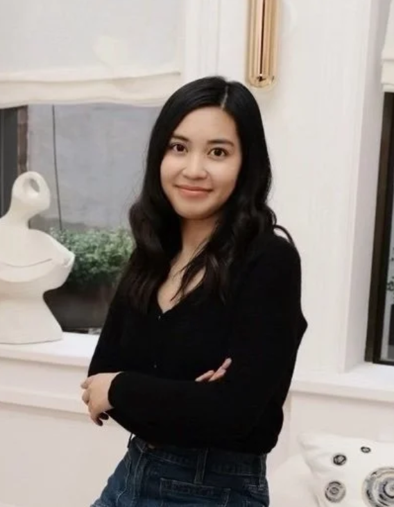 Young woman with long black hair standing indoors, smiling, wearing a black top and jeans, with a decorative duck sculpture and window behind her.