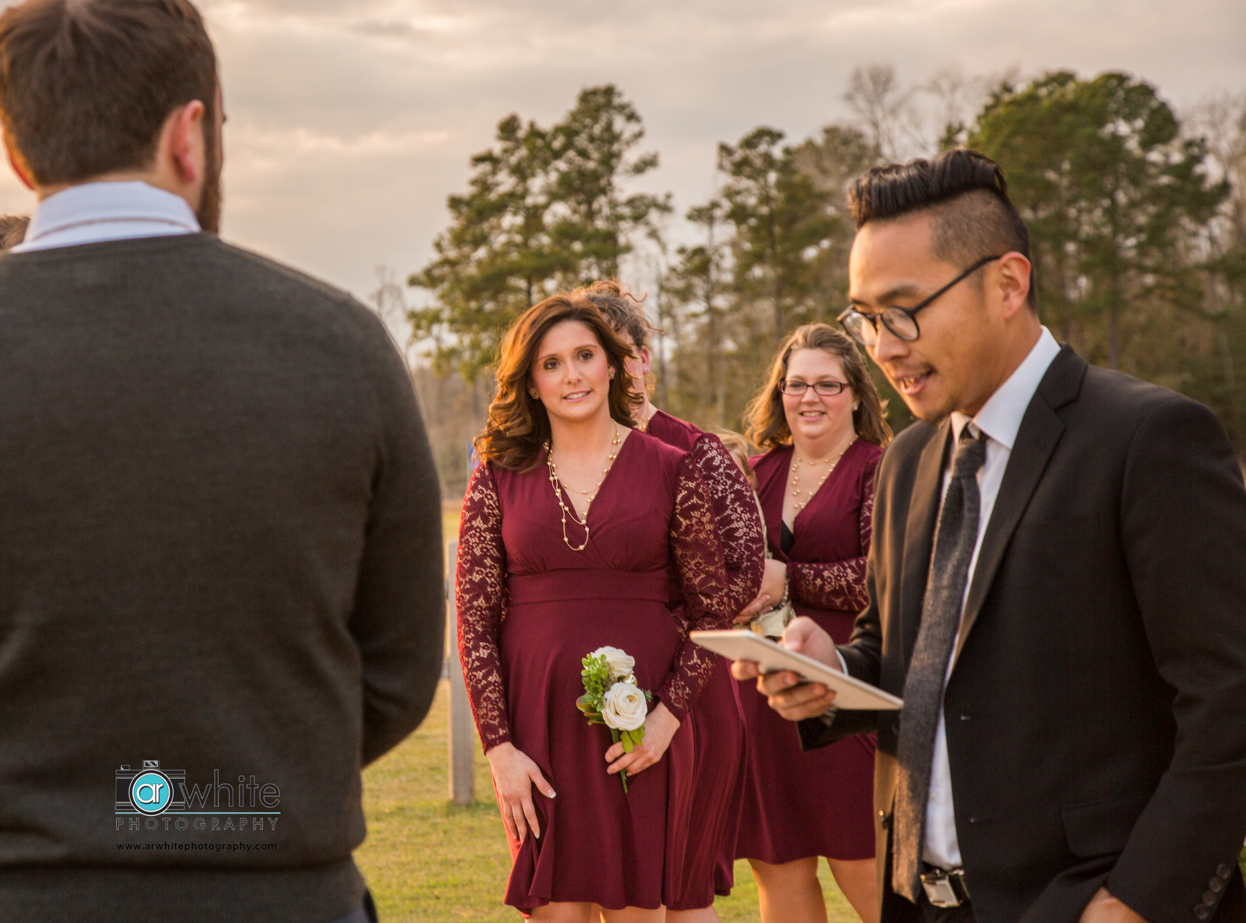 Bridesmaids in maroon dresses with lace sleeves listen to the minister during a wedding reception at a barn wedding venue.