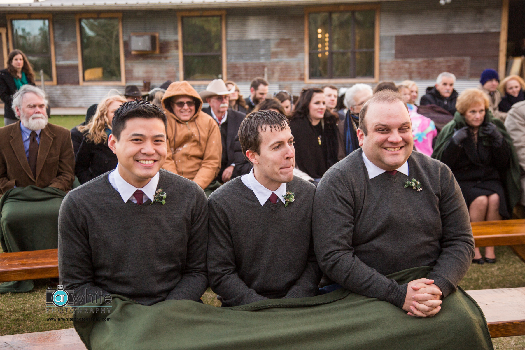 Groomsmen huddle together under a shared blanket during a cold wedding outdoors.