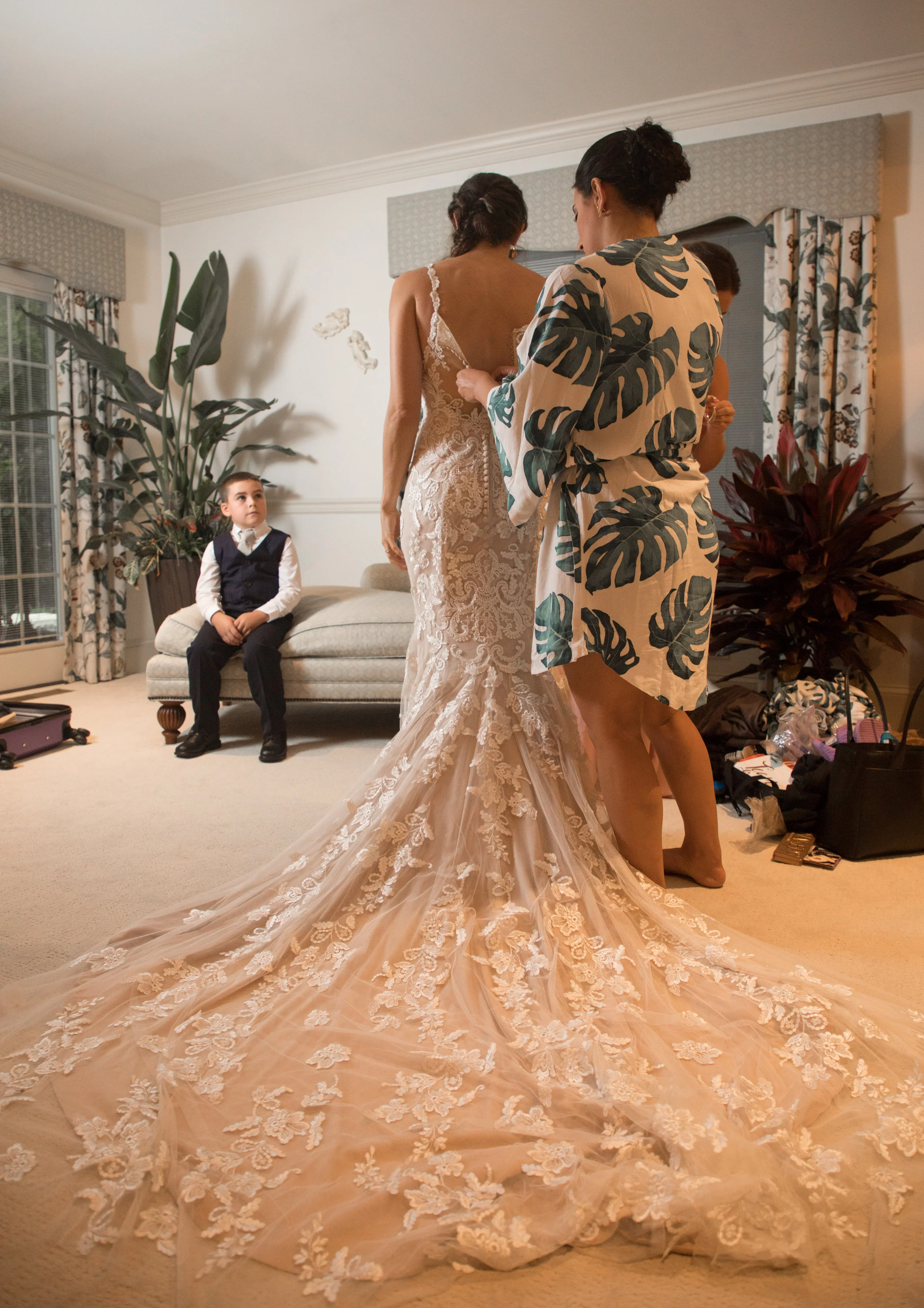 The ring bearer looks admiringly at the bride as her maid of honor buttons the back of her stunning wedding gown.