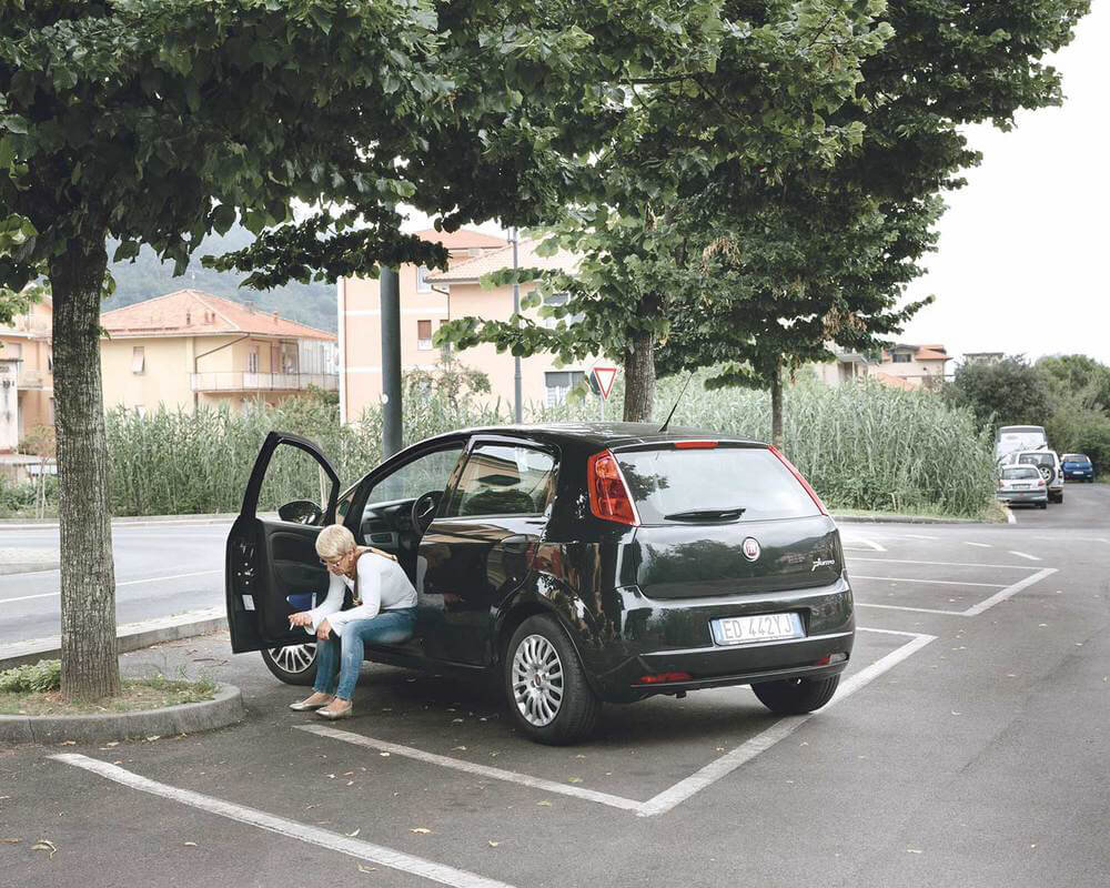 A woman with blonde hair sitting on the edge of a black car, looking at her phone, parked in a lot with trees and buildings in the background.
