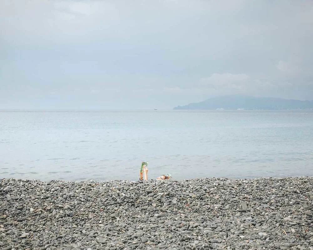 A pebble beach overlooking a calm body of water with a person lying down and a colorful bottle or container nearby. In the background, there's a landmass or island shrouded in fog.