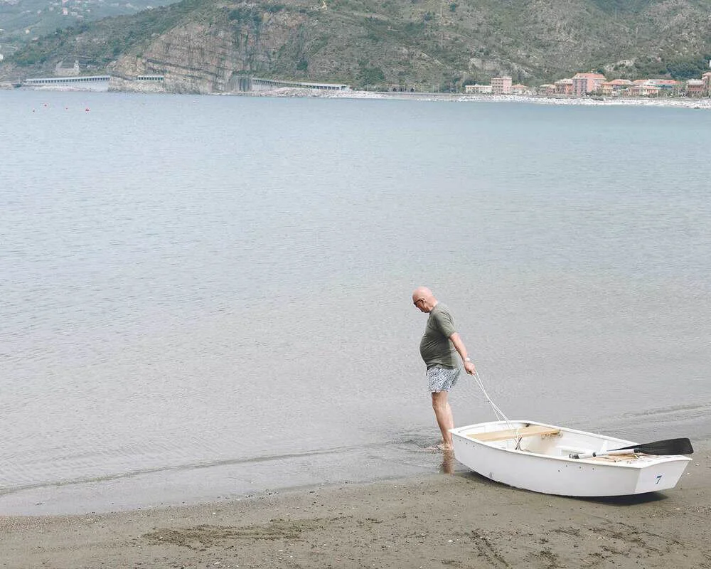 A man with a bald head wearing a gray shirt and patterned shorts standing in shallow water on a beach, holding a small white rowboat with a paddle inside.