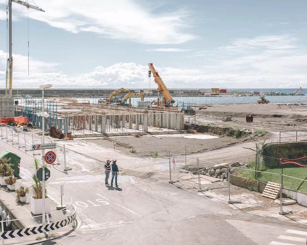 Construction site at a waterfront with cranes and machinery, fencing, and two people walking and talking in the foreground.