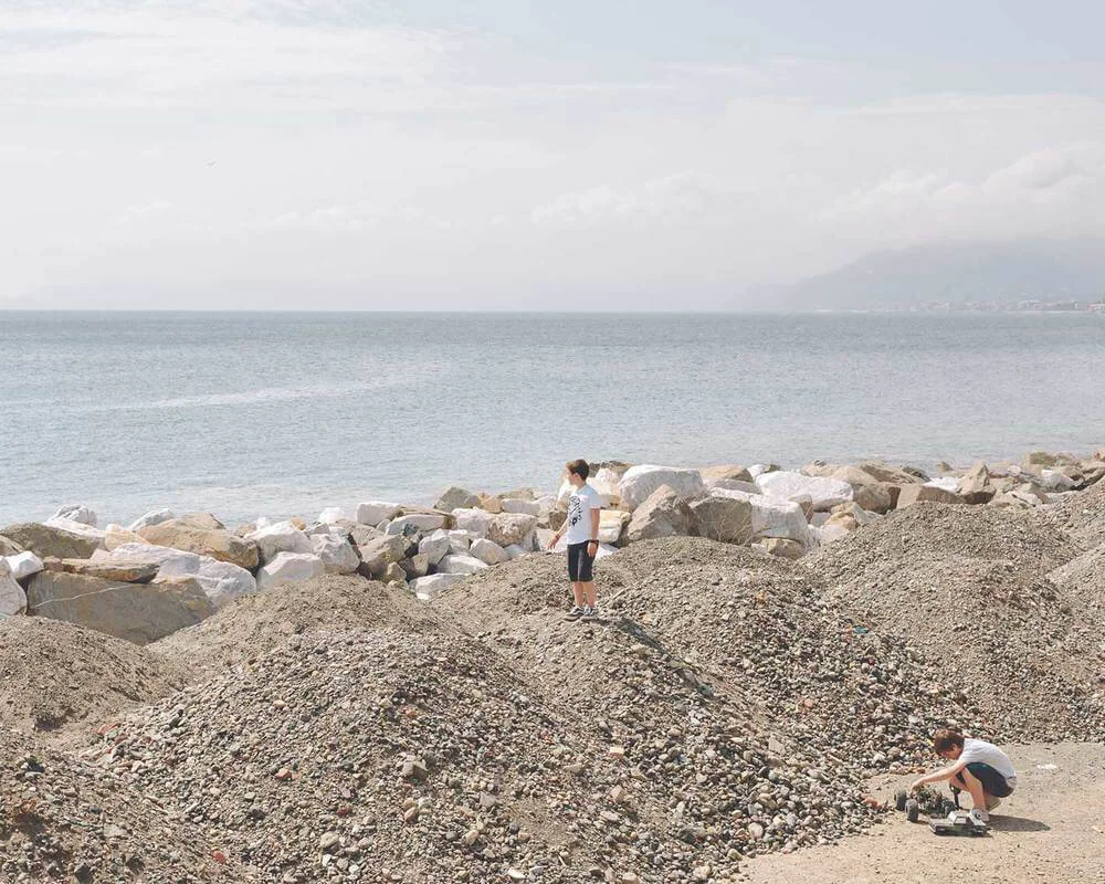 Two boys on a rocky beach, one standing on a mound of gravel and the other crouching near a small toy vehicle.