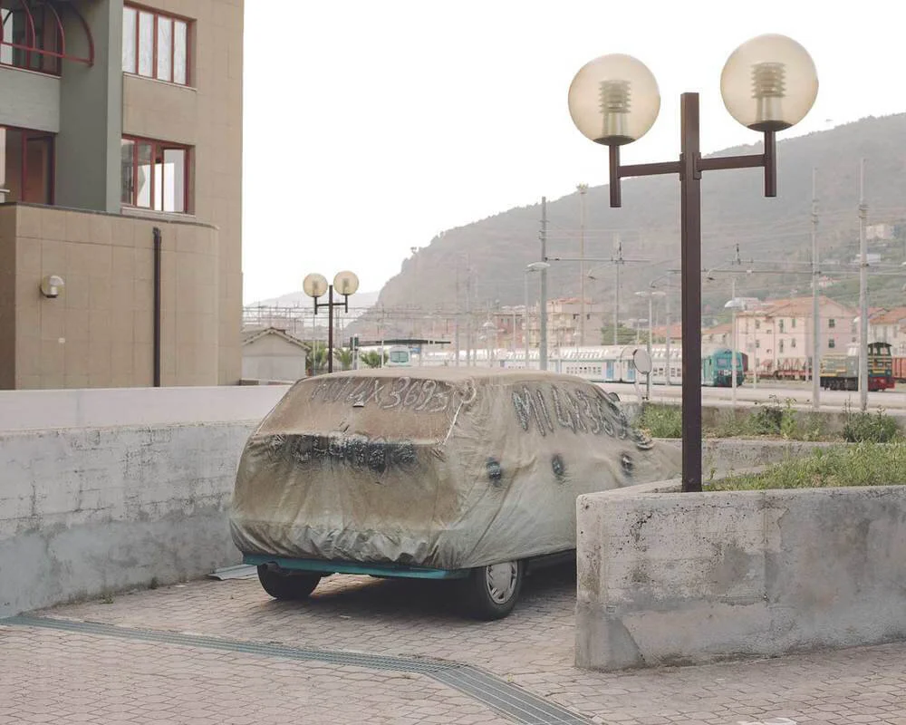 A car covered with a brown tarp, parked on a sidewalk near a concrete wall, with a streetlamp in front of it. In the background, there are buildings, train tracks, and mountains.