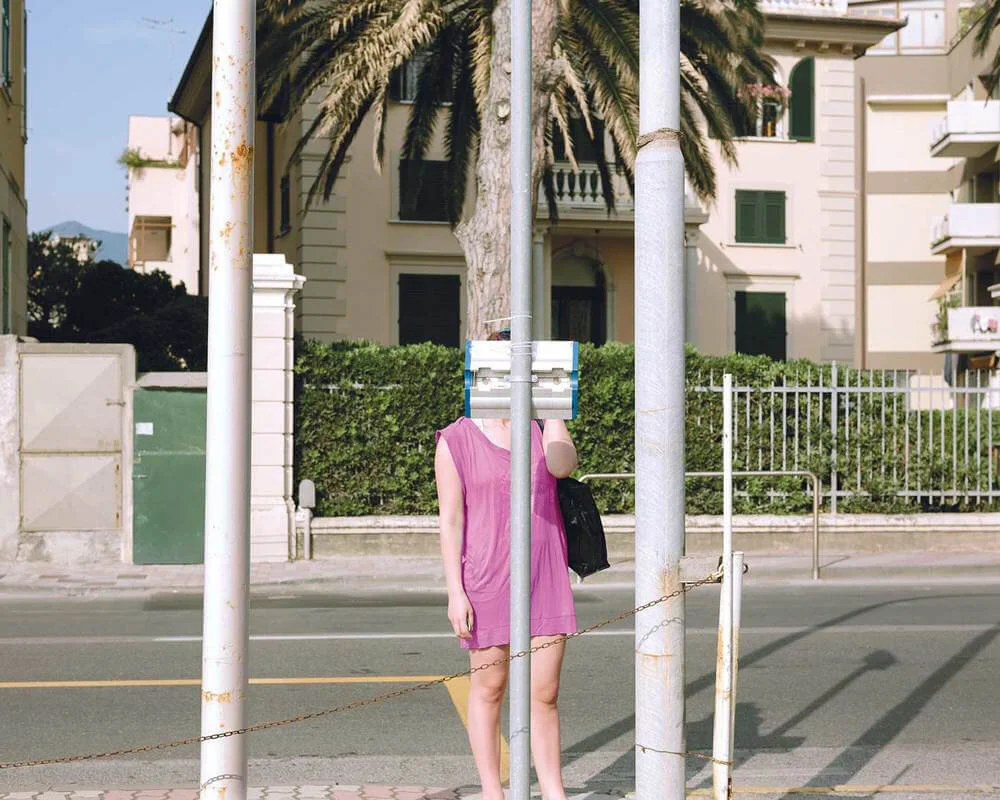 A woman standing behind a street sign in front of a building, with her face obscured by the pole and a bundle of newspapers or magazines. She is wearing a pink dress and holding a black purse.