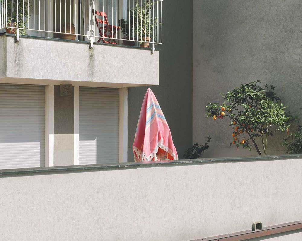 An outdoor balcony with a pink and multicolored striped umbrella, a small orange tree with fruit, and a second floor balcony with red chairs and plants.