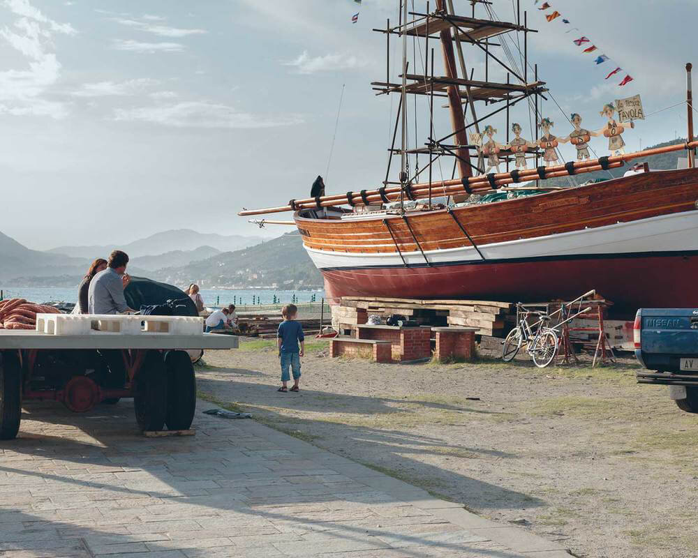 A large wooden ship on land with people nearby, mountains and water in the background, and bicycles and vehicles parked around.