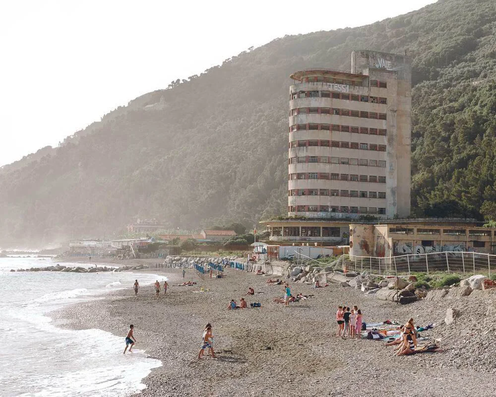A beach scene with people sunbathing and walking, a tall, abandoned hotel building, a mountain in the background, and a partly cloudy sky.