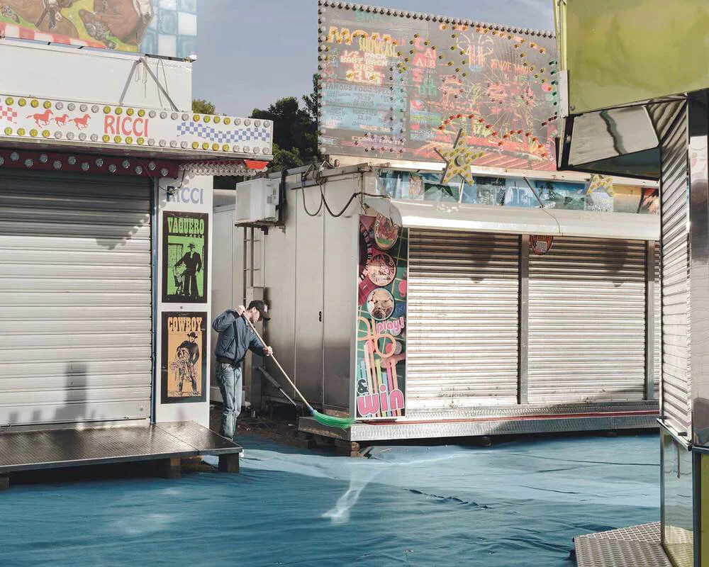 A man watering the ground outside closed carnival game booths with colorful signs and banners, no spectators.