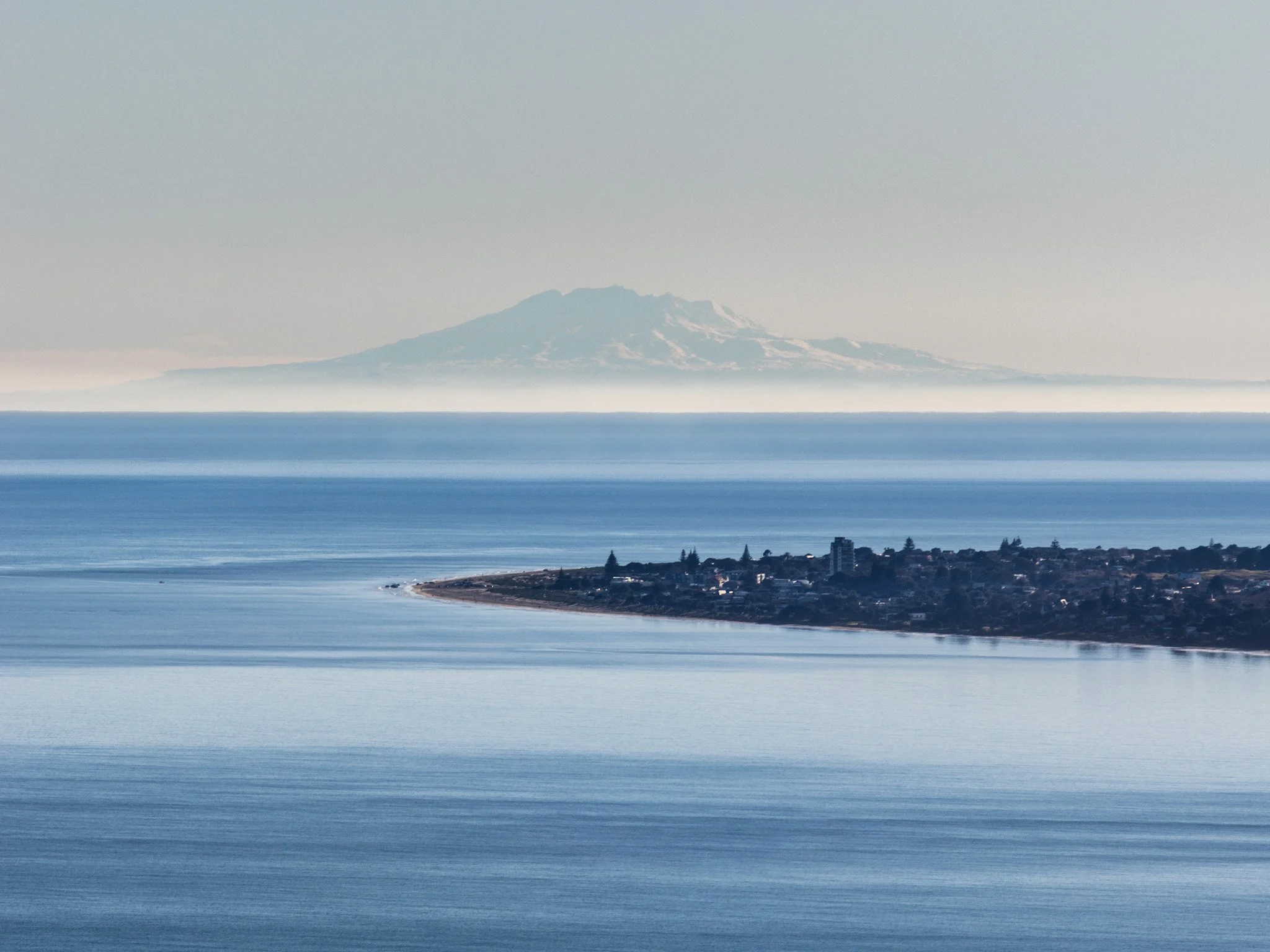 In the lower third, Paraparaumu Beach is surrounded by various tones and textures of the blue ocean. In the background, Mt Ruapehu stands tall, towering above the horizon.
