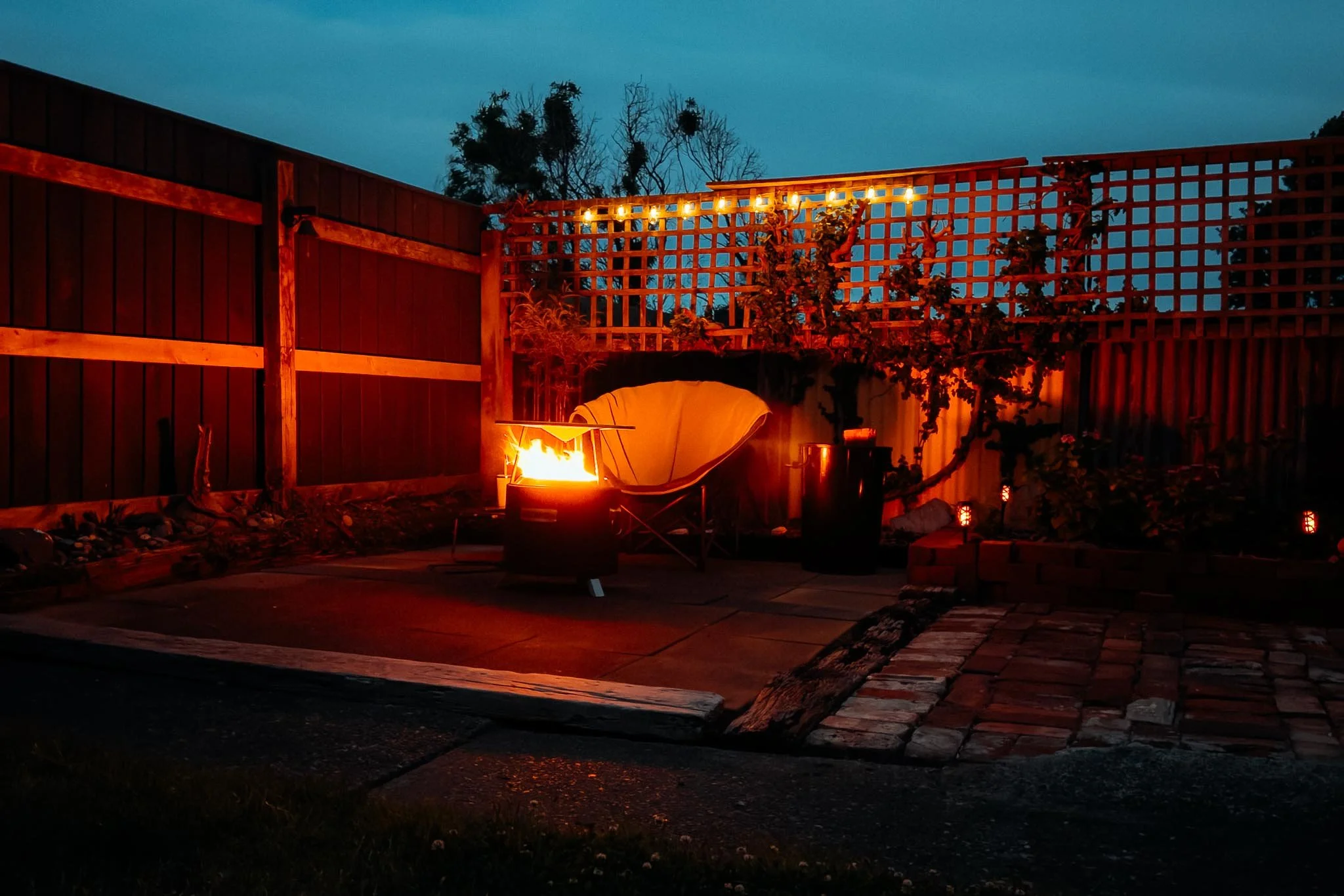 In the corner of a garden, the flames of a Black Wizard fire pit light up the fence and tiles. Small lanterns are around the edge of the garden while a set of solar light bulbs glow along the top of half of one side of the fence.