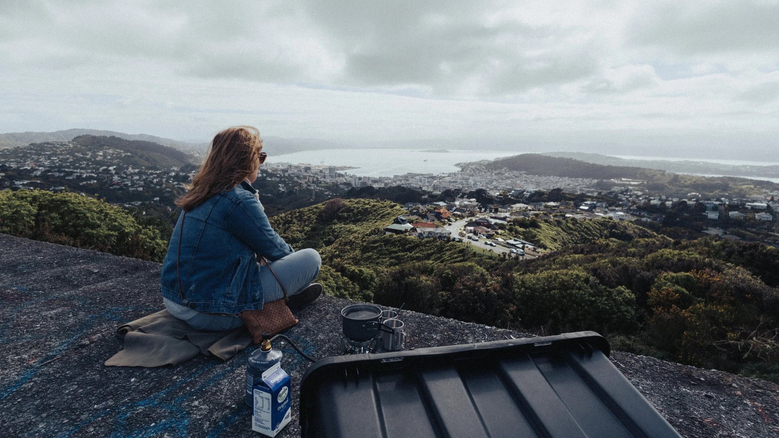 A woman in a blue denim jacket sits on a concrete bunker that sits on top of a hill overlooking Wellington city and harbour under cloudy skies.