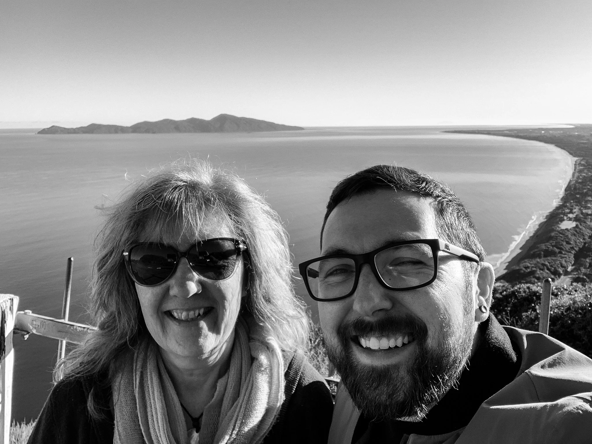 A woman wearing a scarf and sunglasses and a man with a beard wearing glasses take a selfie in front of an island made up of six hills that go from small to larger (left to right) and a coastline that curves out towards the island.