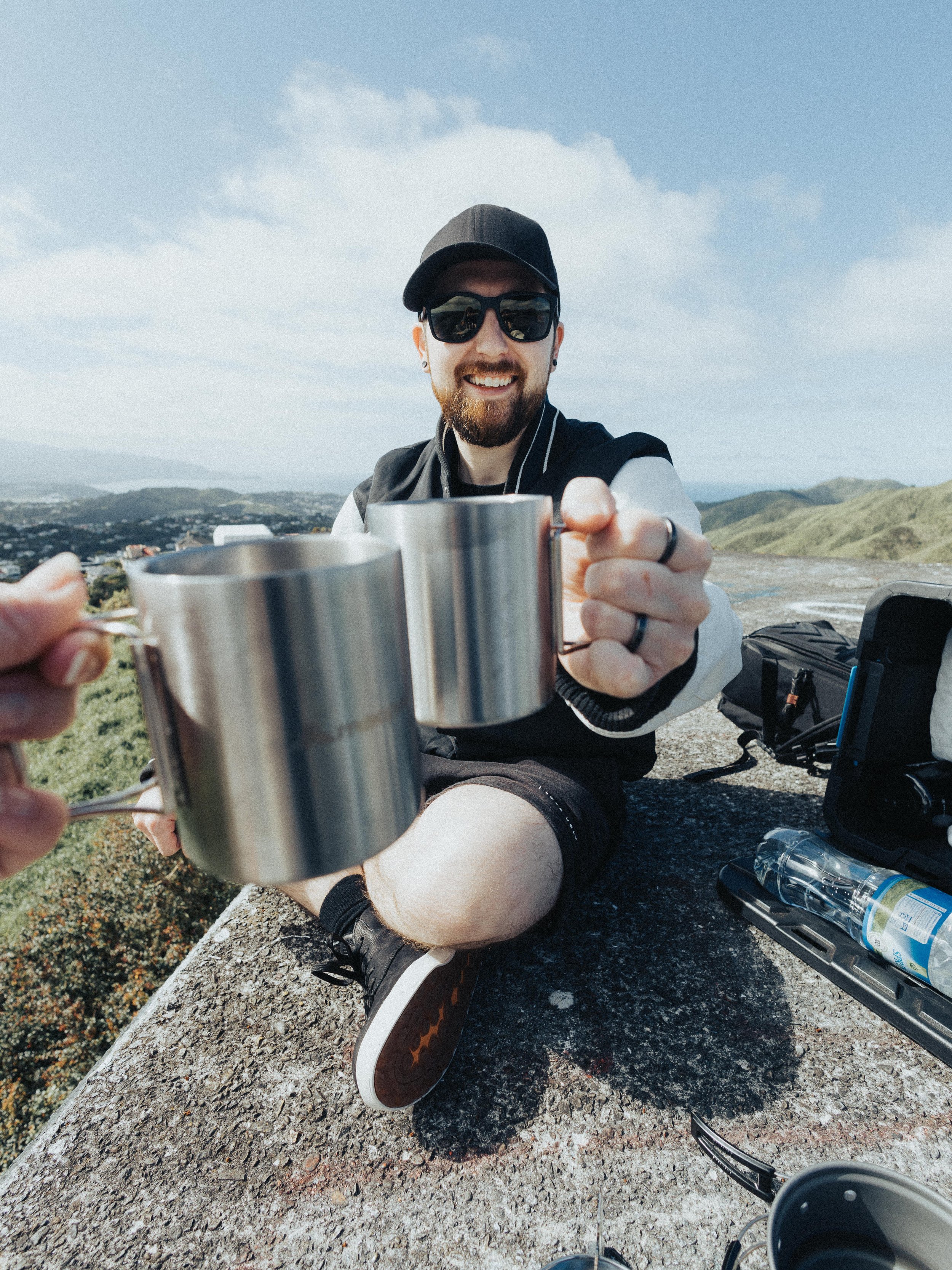 A man wearing a baseball cap, university jacket, shorts and sunglasses, raises a cup of coffee to the camera. In the background is scattered clouds, hills and the ocean on the horizon.