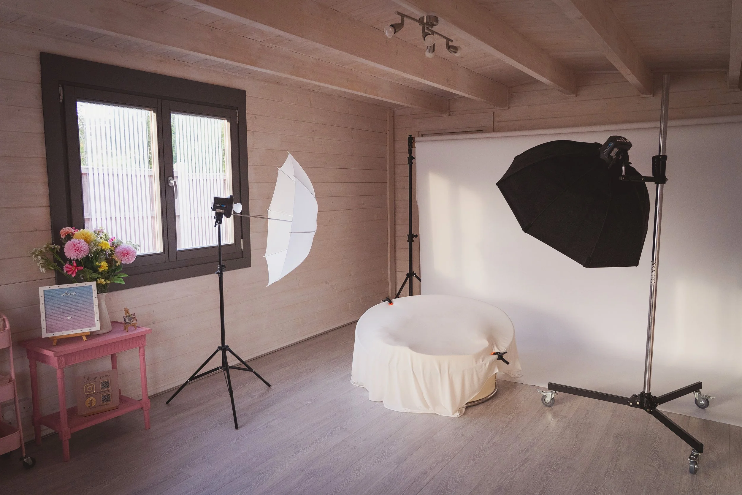 Dani Stoker Photography Studio in Staines. Setup with a white backdrop, studio lights, a white chair covered with cloth, a flowering plant, and a pink side table next to a window.