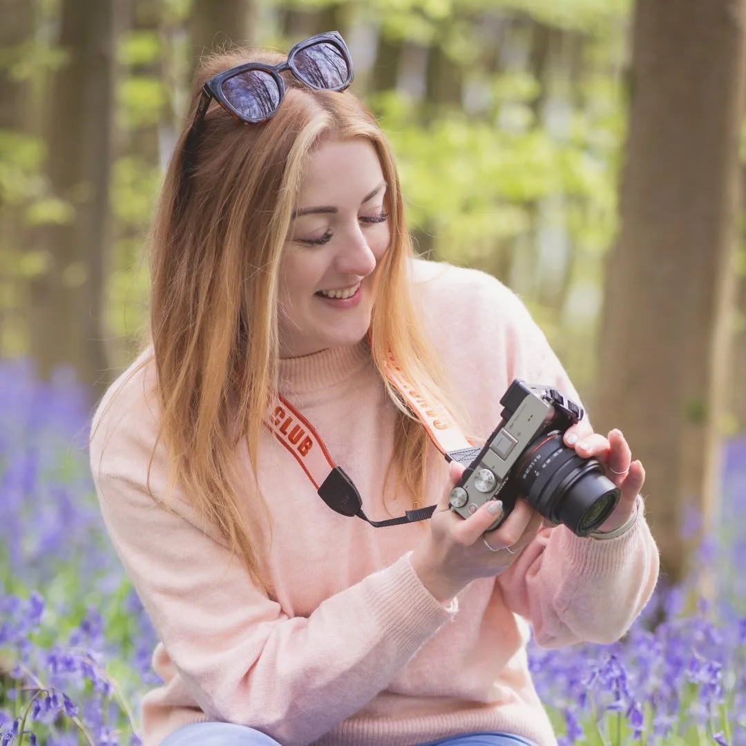 Dani Stoker a Newborn and Cake Smash photographer. A woman with blonde hair and sunglasses on her head, smiling while looking at a camera, outdoors in a wooded area with purple flowers.