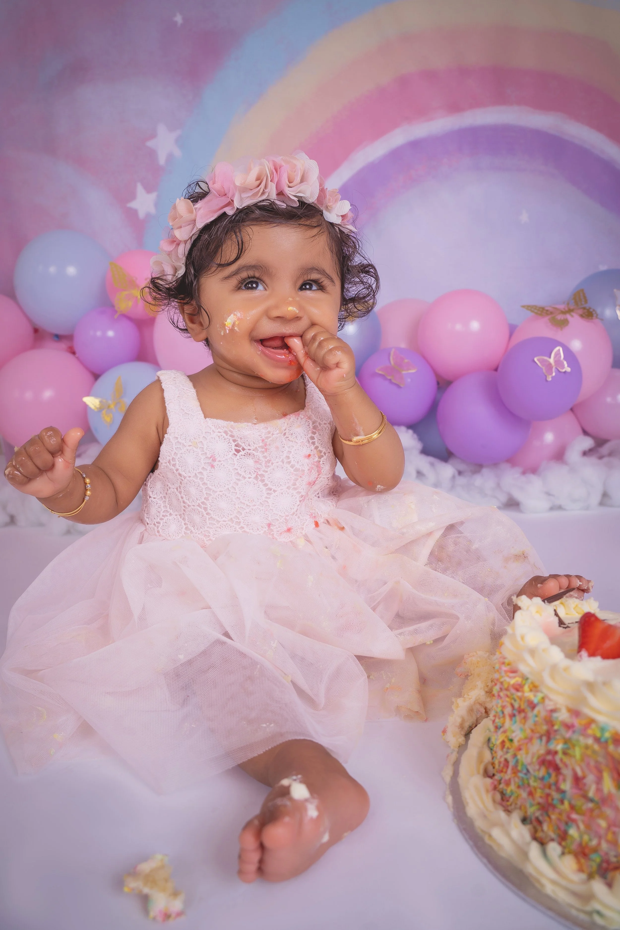 A young girl in a pink dress with a floral headband and gold jewelry, celebrating a birthday with cake and colorful balloons with butterfly decorations, against a pastel rainbow backdrop.
