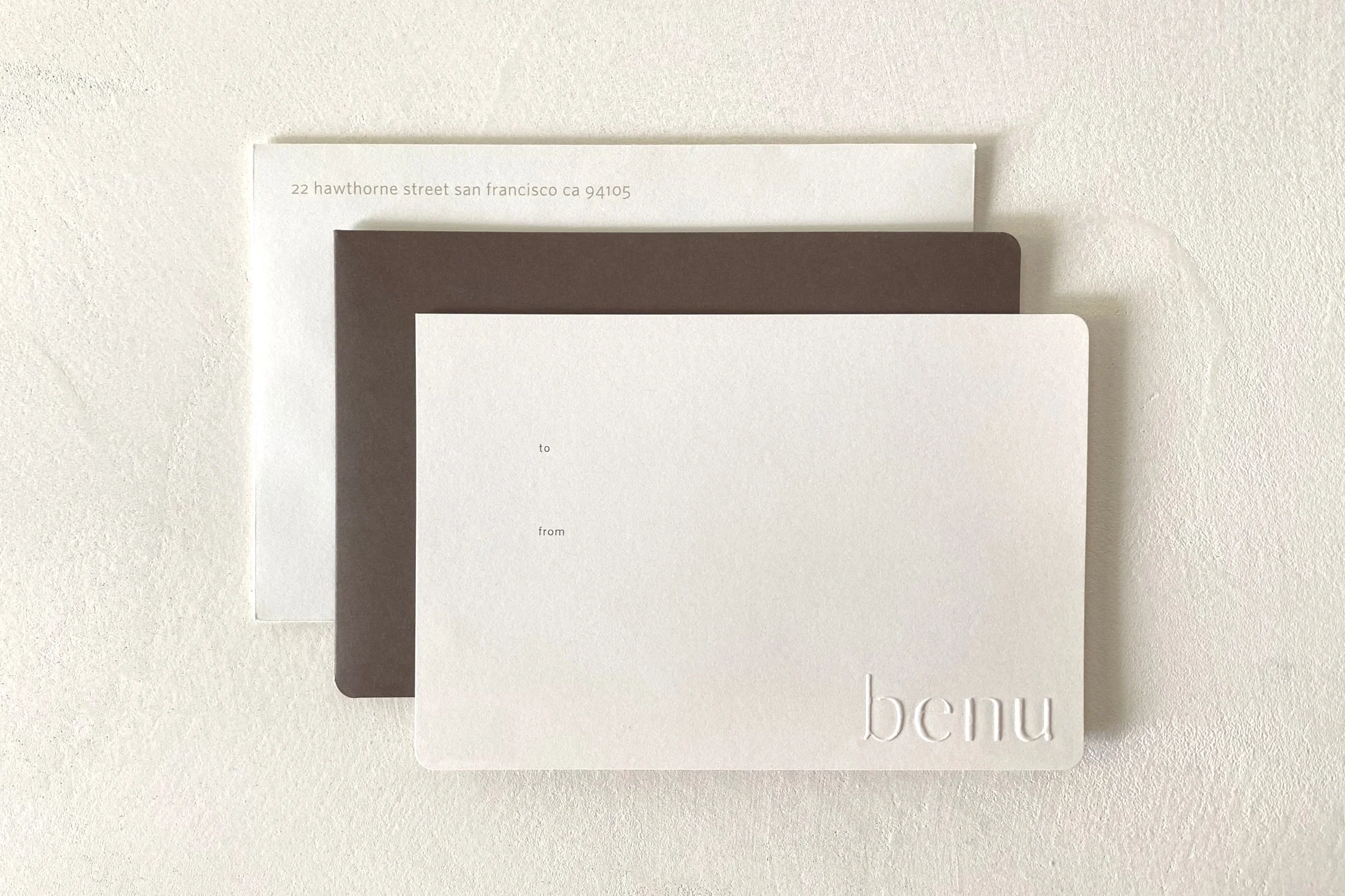 A blank, cream-colored Benu gift certificate stacked atop its deep brown sleeve and a cream envelope.