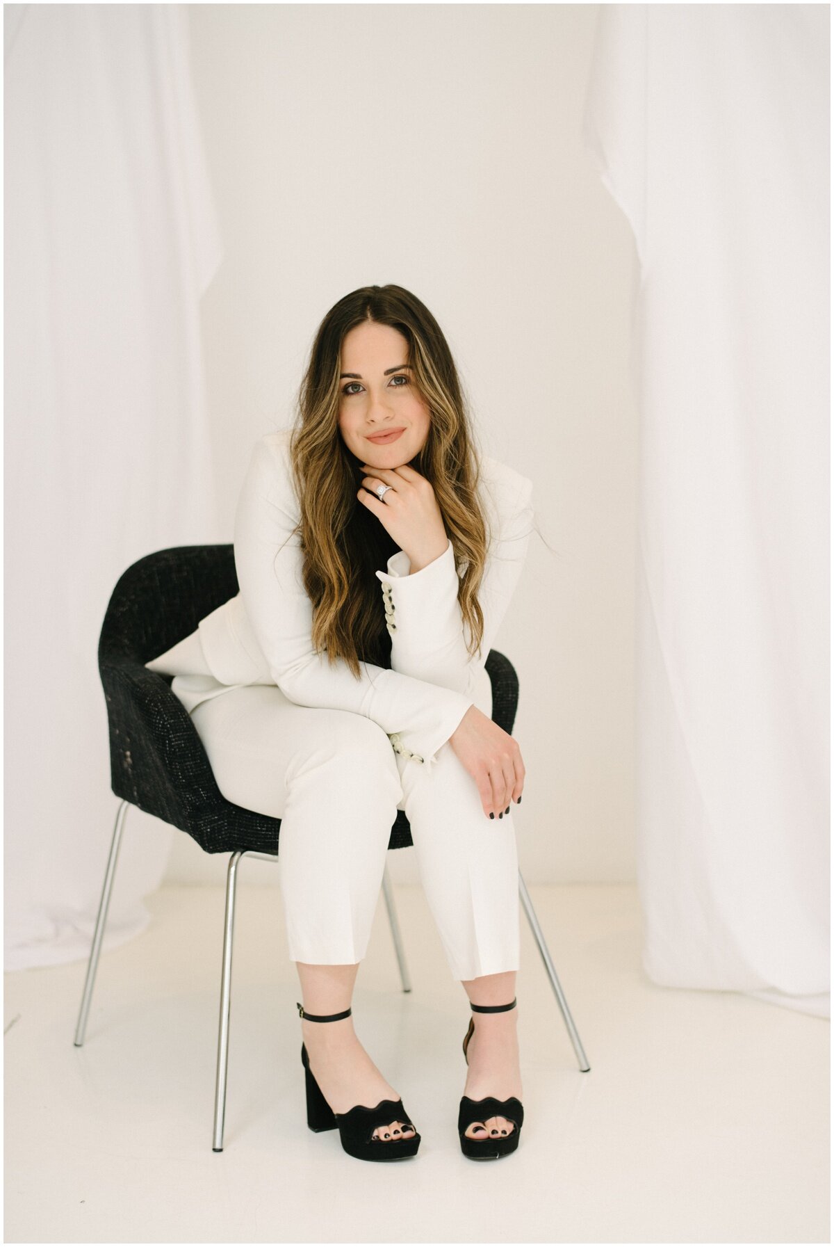 Woman in whit suit with hand on chin, sitting in chair for brand portrait  