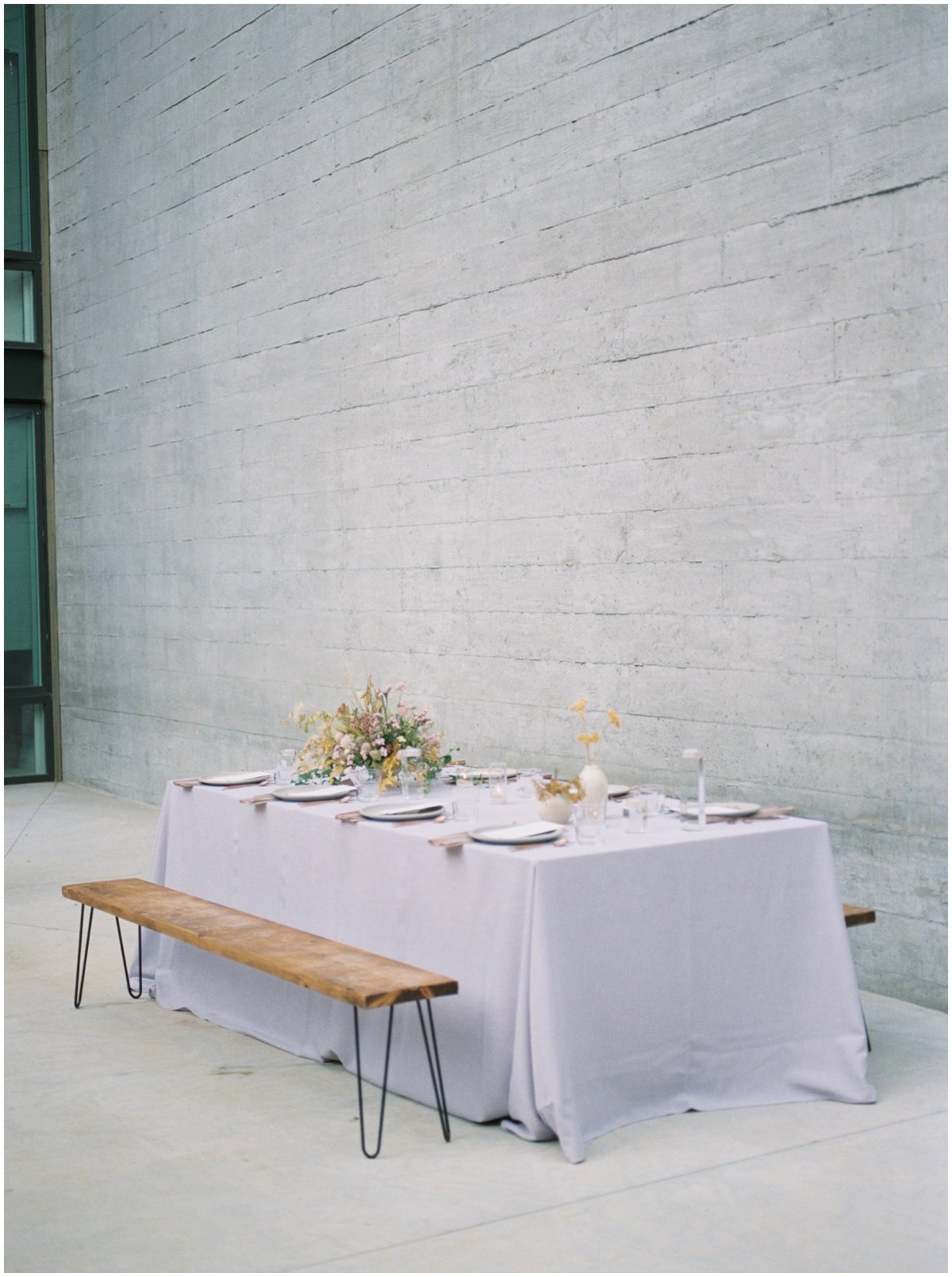 Pale purple table cloth with gold and purple floral arrangements with a wooden bench for seating | NKB Photo