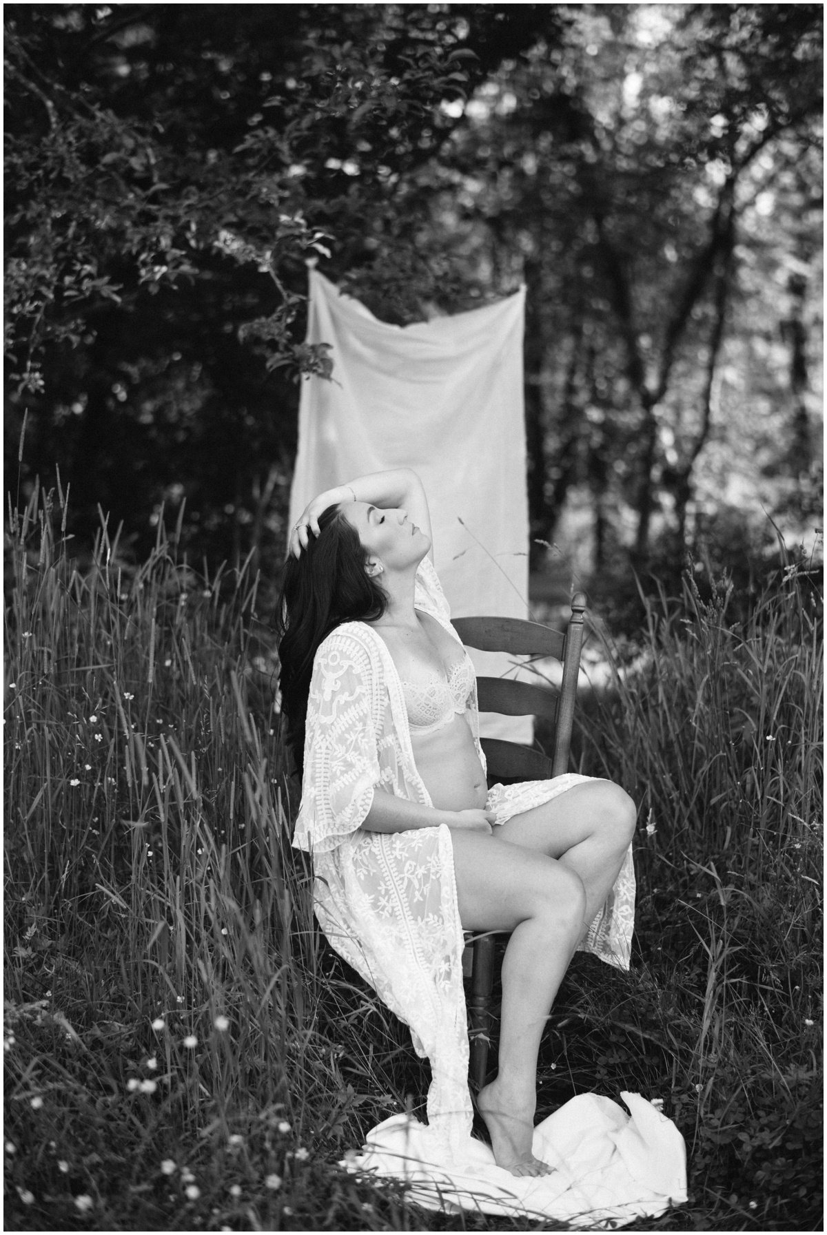 Woman sitting on chair with head thrown back wearing white lace robe during maternity session | NKB Photo