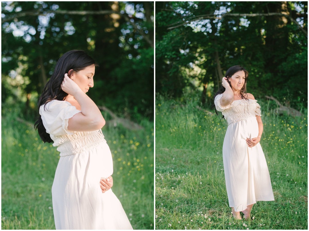 Woman standing in grassy field wearing white dress and cradling baby bump during maternity session | NKB Photo