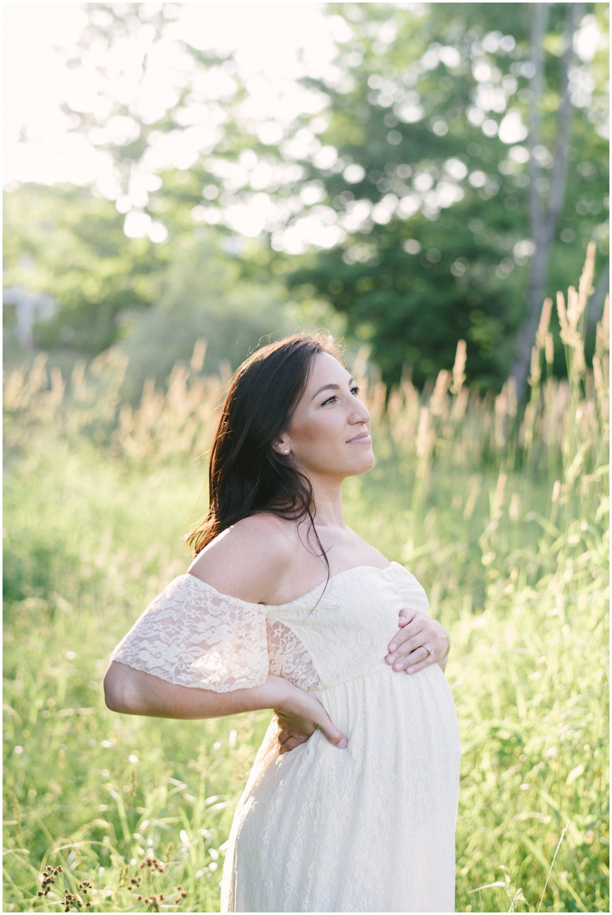 Woman standing in grassy field looking off into the distance while cradling baby bump during maternity session | NKB Photo
