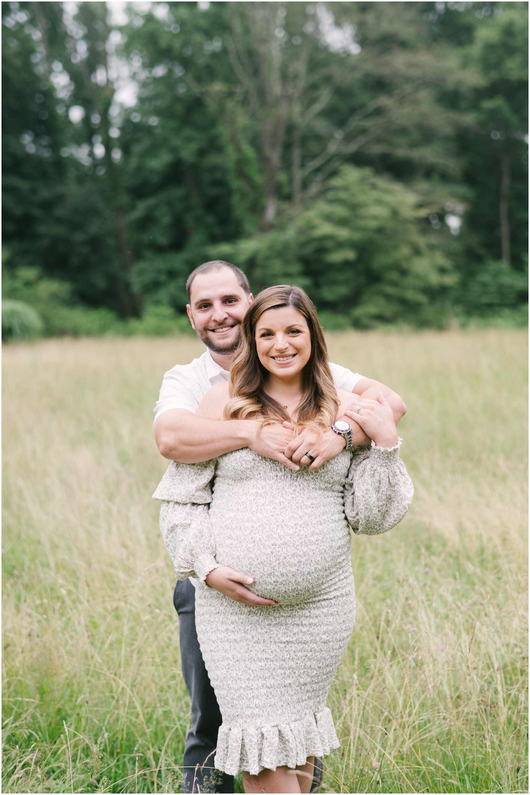 Man standing behind woman while she cradles pregnant belly during maternity session | NKB Photo