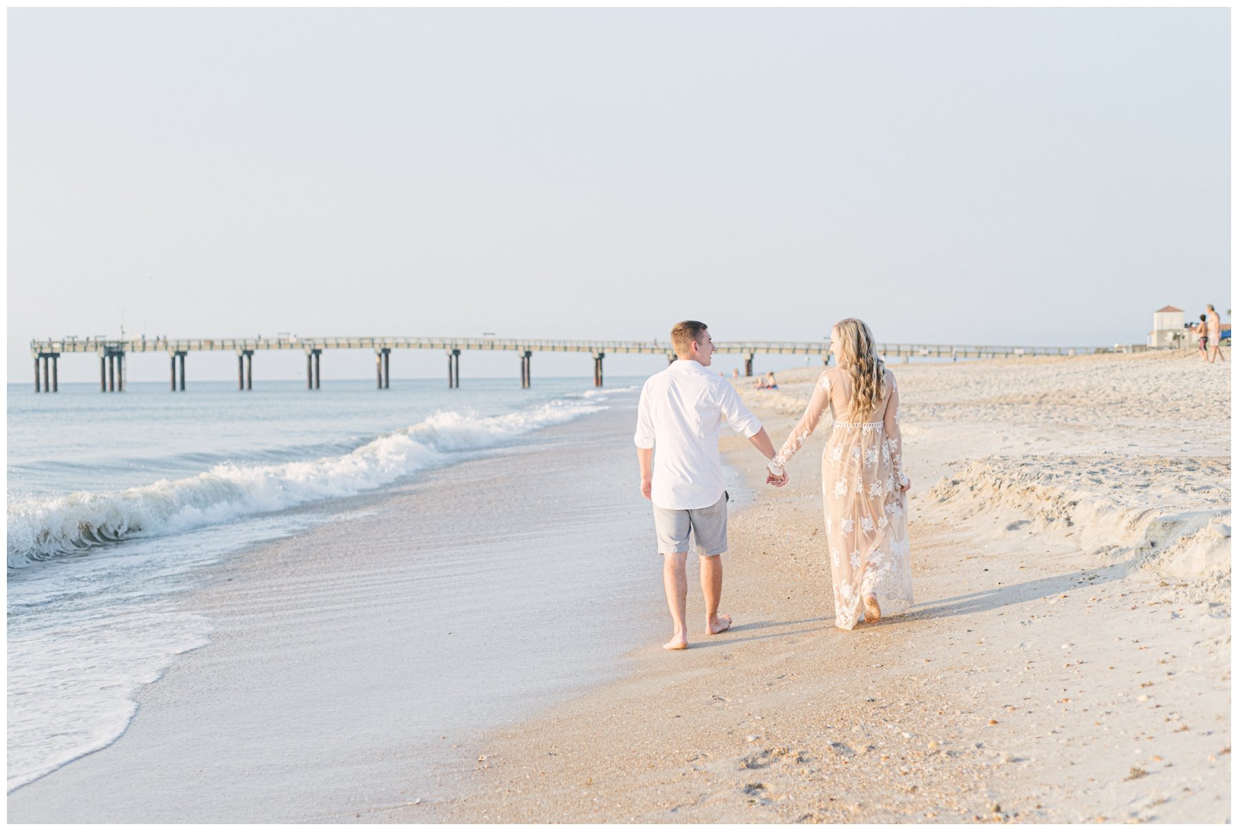 Engaged couple holding hands and walking down beach with pier in background | NKB Photo