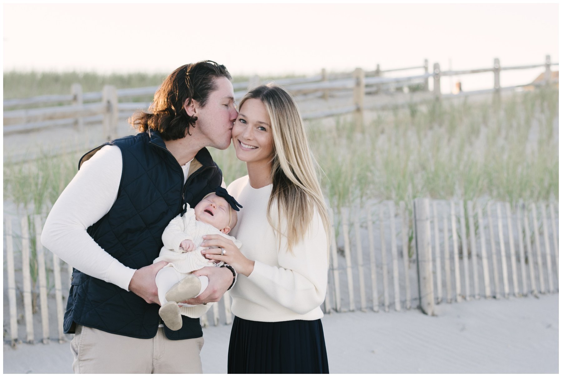 Husband kissing wife while holding newborn during family session with family beach photographer | NKB Photo